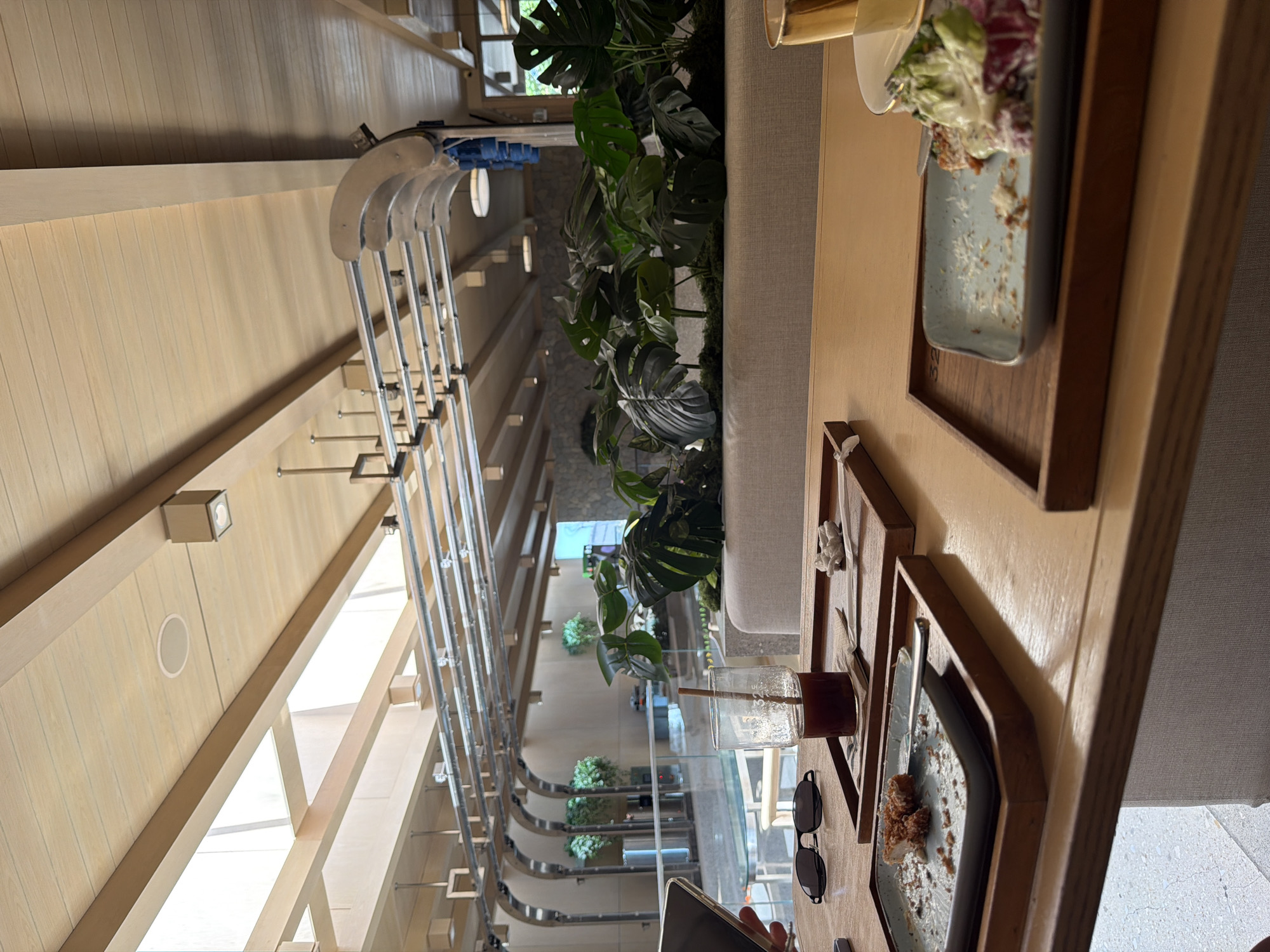This image captures the interior of a modern, airy cafe or restaurant, likely set in a tropical location such as North Kuta, Indonesia, given the context and visible flora. The architecture features light-colored wooden slatted walls and ceilings, creating a bright and contemporary aesthetic. Large windows or skylights allow ample natural light, suggesting it is daytime.
In the foreground, a wooden table is set with items indicating a casual dining experience. A glass with a dark beverage (possibly iced coffee or tea) and a straw is partially consumed. A pair of black sunglasses rests beside it. A light blue rectangular plate, mostly empty with some food remnants (crumbs or dressing) on a wooden tray, suggests a recently finished dish. Another wooden tray holds a small white coral piece. A hand holding a smartphone is partially visible on the table, indicating a person is present, though no faces are seen. Further back on the table, another plate contains a salad with green and red leaves.
A long, light-colored upholstered bench or sofa runs through the midground. Behind it, a significant feature is a planter filled with large, lush green tropical plants, likely Monstera, which adds a vibrant, natural element to the interior design.
The background showcases the cafe's structural design, including exposed silver metal beams or tracks running along the high ceiling, possibly for lighting or decorative purposes. Additional recessed lighting fixtures and small square spotlights are visible on the ceiling. More greenery can be seen in the distance, contributing to the serene and open atmosphere. No discernible text is visible in the image.