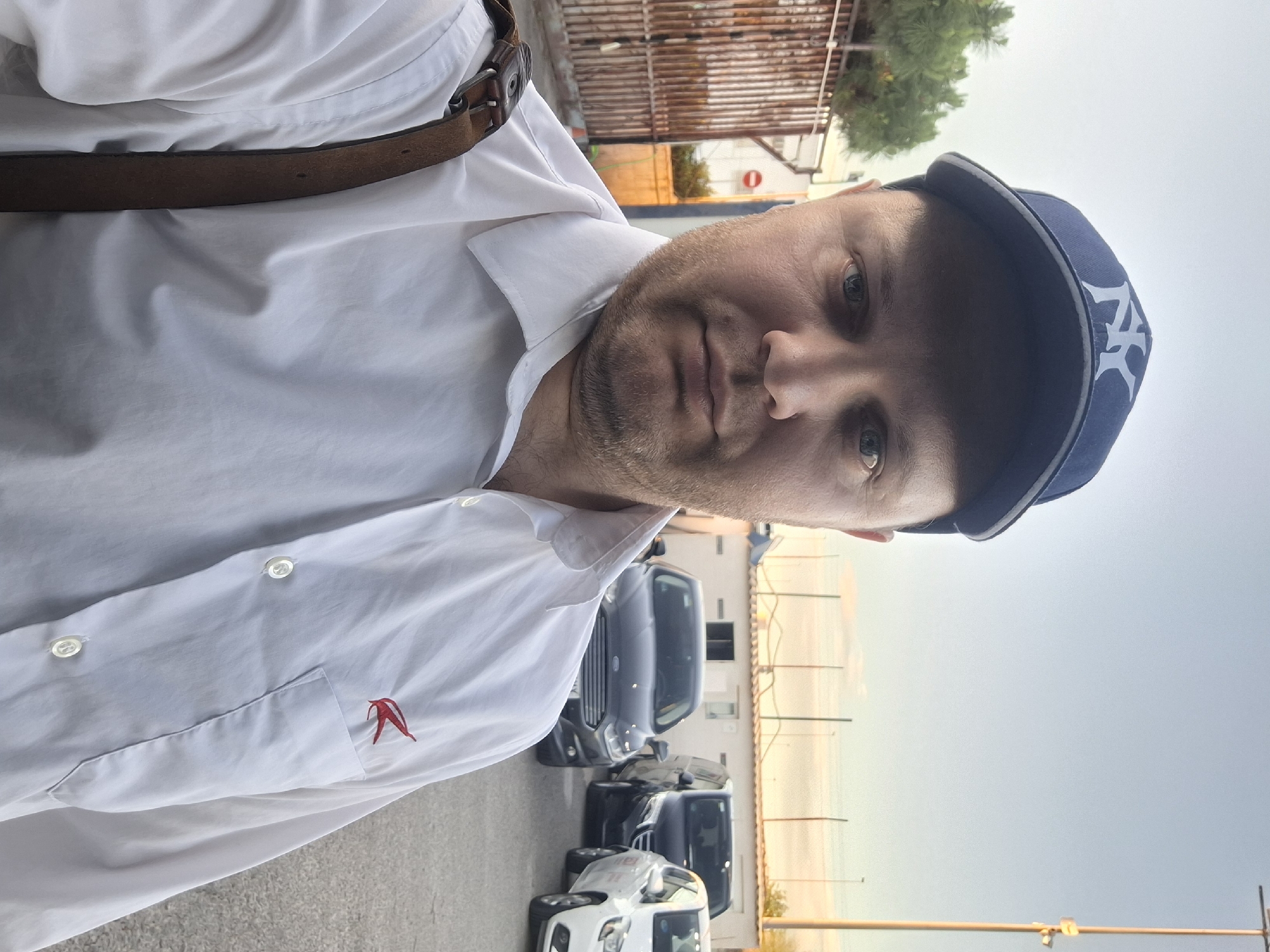 A man wearing a white collared shirt, a brown leather belt, and a dark blue baseball cap with a white "NY" logo is taking a selfie. He appears to be standing in a parking lot in the city of Quart de Poblet, Spain. There are several cars parked behind him. A chain-link fence and a building can be seen in the background. The sky is overcast.