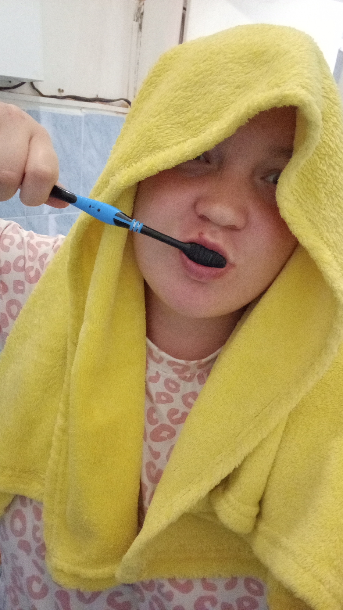 A person with fair skin, wearing a yellow hooded towel, is brushing their teeth with a black and blue toothbrush. The person is wearing a pink and white spotted shirt. The location appears to be a bathroom with blue-tiled walls. The city is unknown.