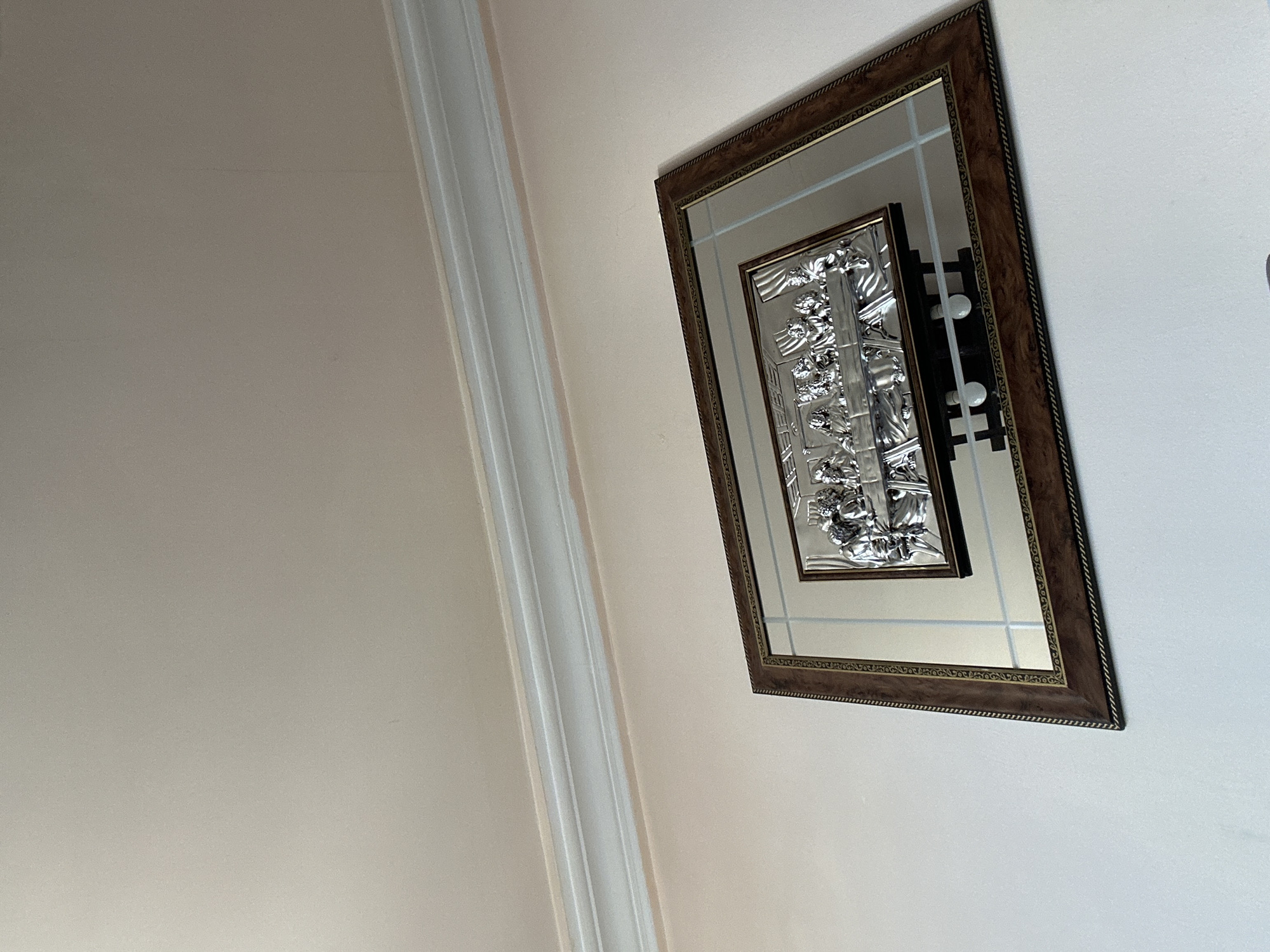 An indoor scene features a decorative framed artwork mounted on a light-colored wall. The frame is rectangular, oriented vertically, and has a dark brown, possibly wooden, border with intricate detailing along its edges. Inside this frame is a mirrored surface. Within the larger mirror, a smaller, dark-framed piece of art is centered. This inner artwork is a silver-toned, embossed, or sculptural relief, displaying ornate patterns and possibly figures, hinting at a classical or elaborate style.
Reflected in the main mirror are sections of a light-colored wall or ceiling, along with some linear structural elements. More notably, the mirrored surface of the inner artwork clearly reflects what appears to be part of a light fixture, showing two prominent circular light bulbs or similar white objects mounted on a dark, vertical support.
To the left of the framed artwork, a smooth, light beige or off-white wall extends. A white or off-white ceiling molding or architectural trim runs vertically along the left edge of the frame, separating the wall plane from what might be a ceiling or another wall. The image is captured from a slightly upward and angled perspective, causing the frame to appear askew. The lighting is soft and diffused, creating no harsh shadows. No activity is taking place, and no text is visible.
