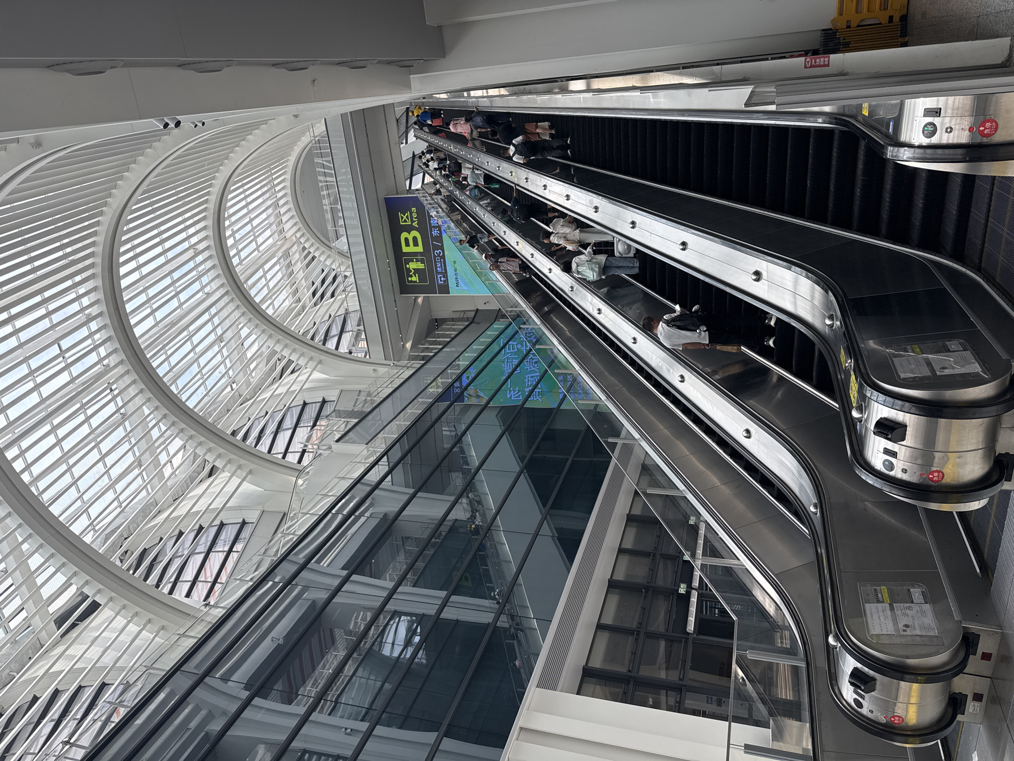 The image shows an interior view of a modern building, likely a transportation hub like an airport or train station, with a distinctive arched glass ceiling. Multiple escalators are visible, some ascending and some descending, with many people using them. The escalators are stainless steel with black treads. People of various ages and genders are dressed in casual clothing, some carrying luggage. A prominent sign is visible in the upper right portion of the image, featuring a "B Area" designation along with Chinese characters and an icon depicting people entering. The architecture of the building is characterized by large glass panels and white structural elements forming the arches of the ceiling. The lighting appears to be natural, coming from the glass roof, and supplemented by interior lighting. The overall scene depicts a busy but orderly environment with people traveling. Based on the provided context, this scene is located in Baiyun District, China.