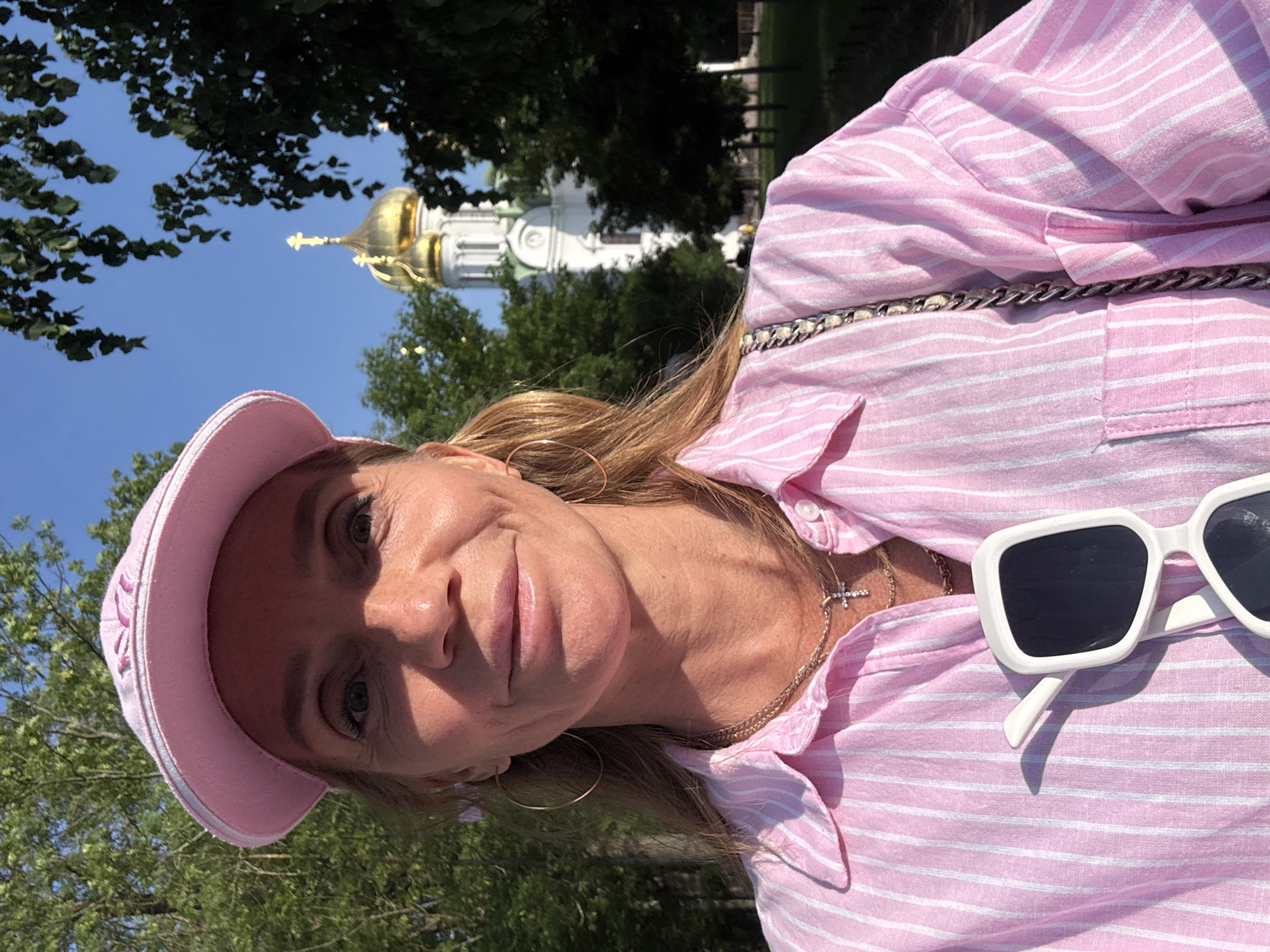 A woman in a pink and white striped collared shirt and a pink baseball cap is taking a selfie outdoors. She is wearing gold hoop earrings and a gold necklace with a cross pendant. She has white sunglasses hanging from the front of her shirt. In the background, a white church with golden domes is visible through trees. The location is the city of Pushkin, Russia.