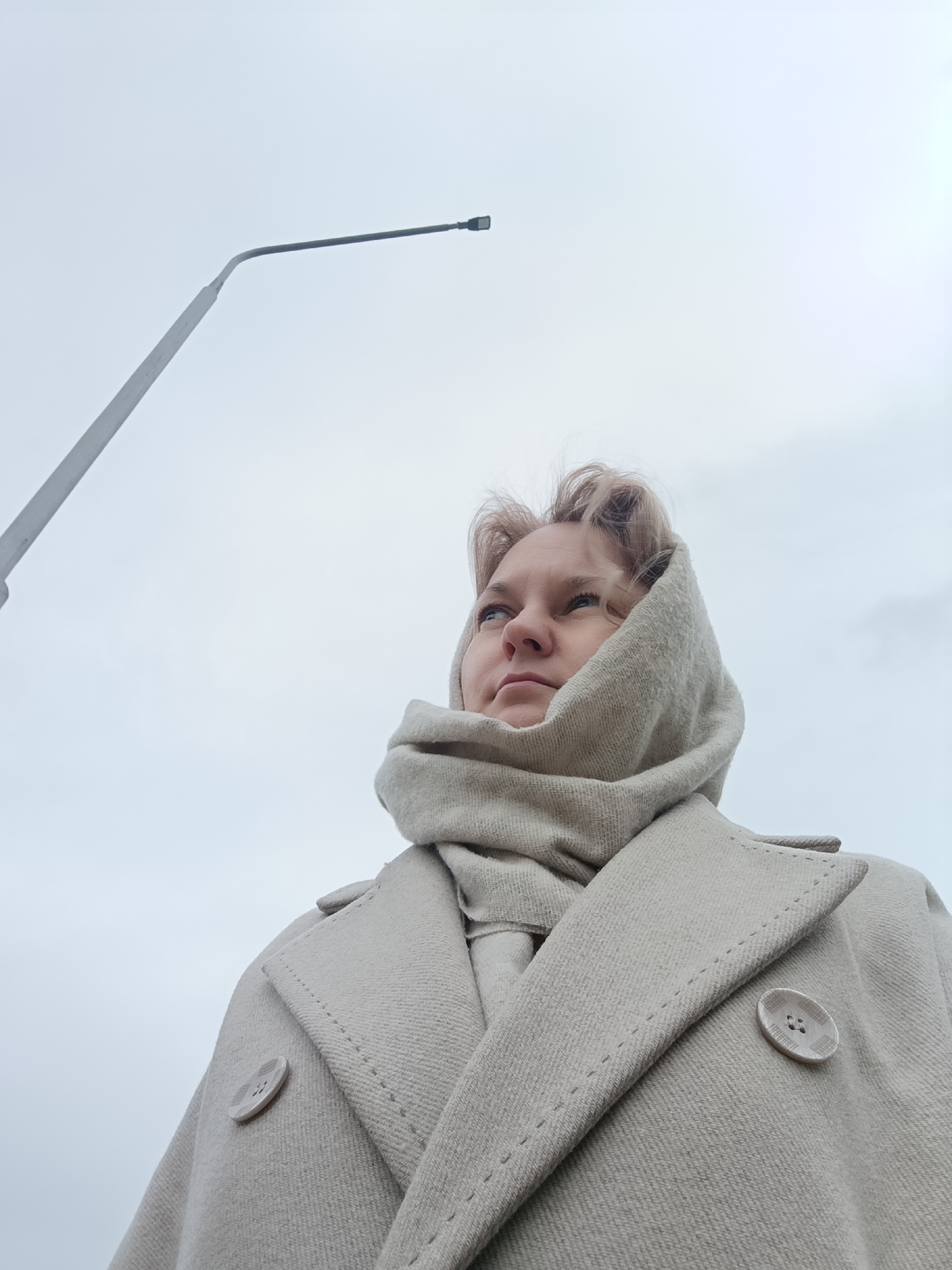 A low-angle outdoor shot features a woman looking upwards and to her left, against a light grey, overcast sky. The woman has light blonde or light brown hair and is wearing a thick, light beige or cream-colored coat with prominent, round, patterned buttons on the lapel. A matching scarf or hood is wrapped around her head and neck, covering her ears. Her expression is pensive and observant, as she gazes towards something out of frame.
In the upper left portion of the image, a dark grey streetlight pole extends diagonally upwards from the bottom edge, then bends to the left as a horizontal arm, ending in a dark fixture. The background consists entirely of the pale, cloudy sky, suggesting a cool or cold day without direct sunlight. The scene is likely set in an urban or suburban outdoor environment, consistent with the provided location context of Markovsky, Russia. No visible text is present in the image.