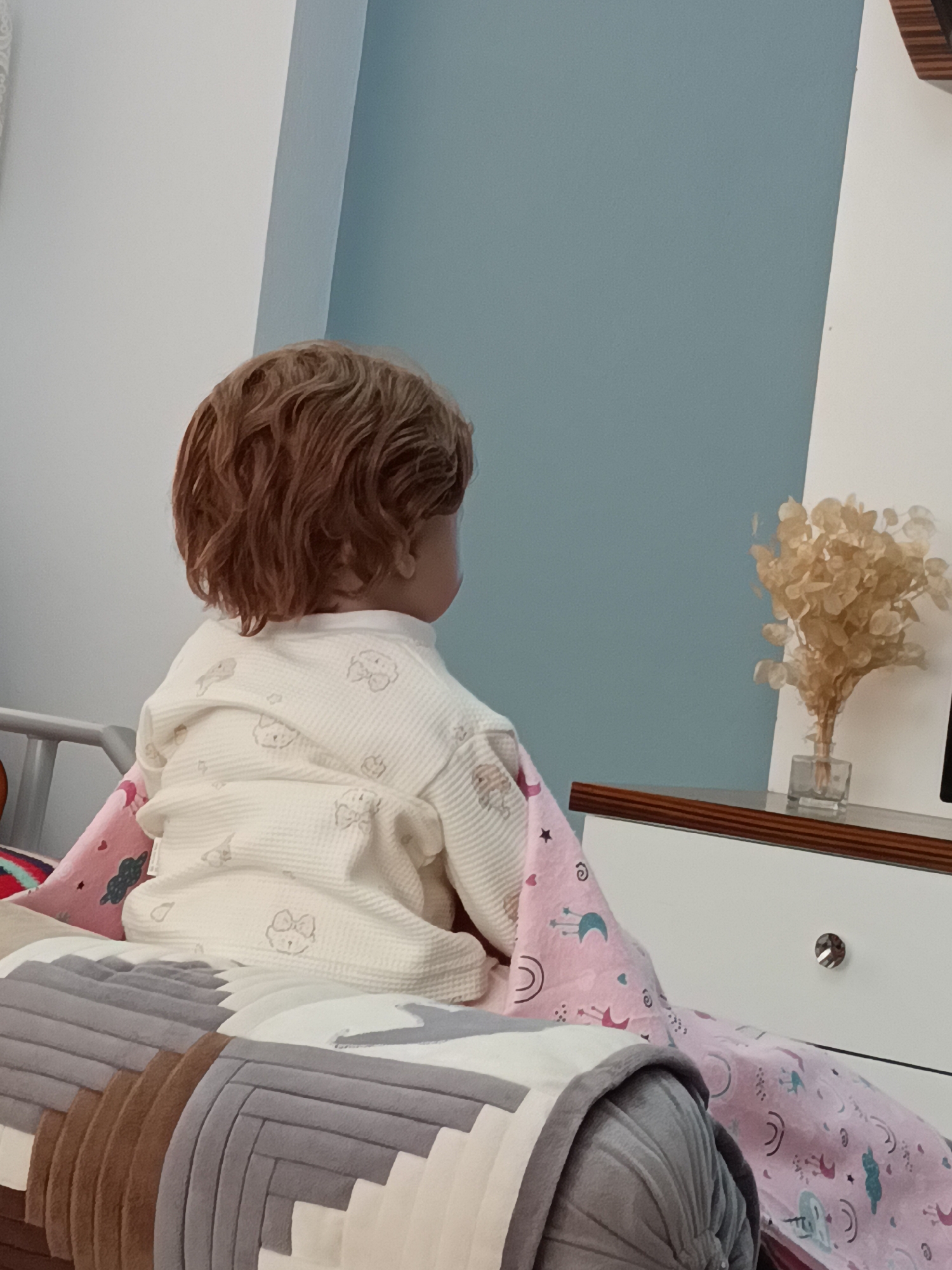 The media file shows the back view of a doll sitting on a bed. The doll has wavy, reddish-brown hair and is wearing a light-colored pajama set with animal prints. A pink blanket with celestial motifs is draped over the doll. In the background, a light blue wall meets a white wall, with a wooden drawer unit positioned between them. A small glass vase containing dried flowers sits on top of the drawer unit. The scene appears to be indoors, possibly a bedroom, with soft lighting suggesting it is daytime. There is no visible text in the image. The overall impression is one of a quiet, domestic setting.