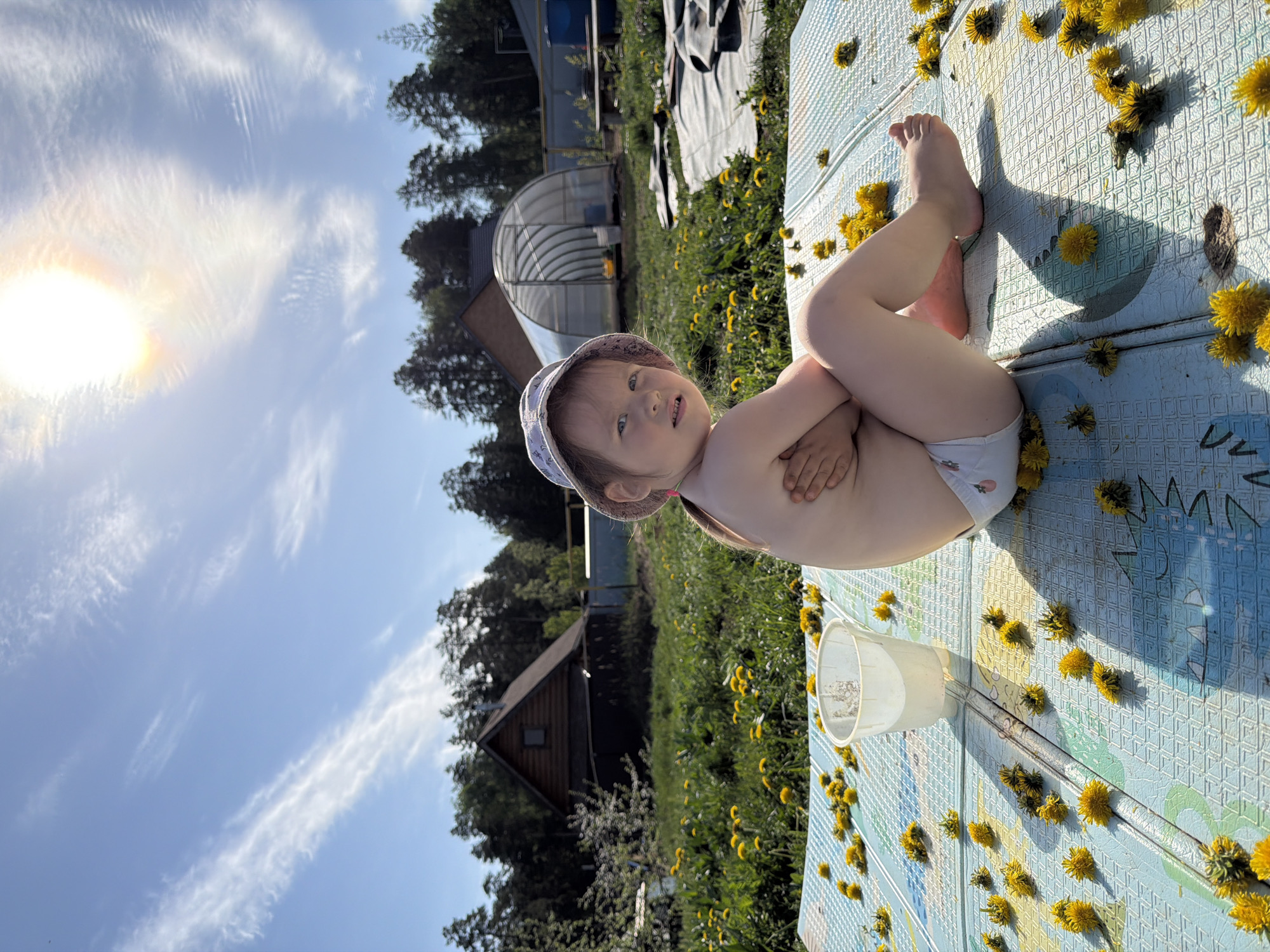 A young child wearing a sunhat and a diaper sits on a patterned, light-blue mat scattered with yellow dandelion flowers in a grassy, outdoor setting. The child is positioned facing the viewer with their legs crossed and arms folded. Beside the child is a clear plastic cup. In the background are various buildings, including a wooden house and a greenhouse, surrounded by trees and vegetation. The sky is visible with some wispy clouds and a bright sun. This scene depicts the outdoor area of a house, presumably located in Chaikovsky, Russia.