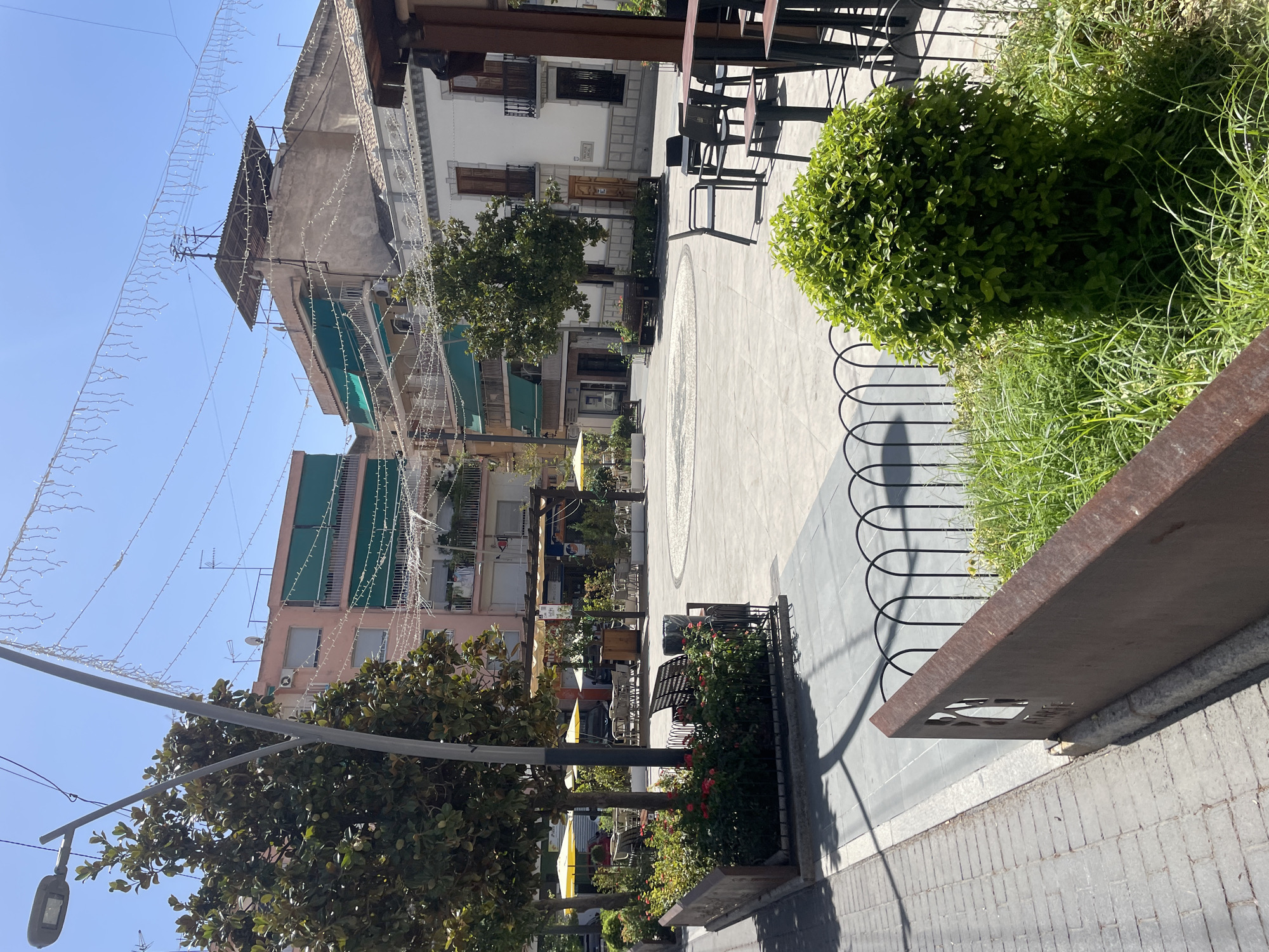 This is a daytime, eye-level, outdoor photograph of a paved plaza in Atarfe, Spain. The plaza is lined with trees and includes outdoor seating areas for cafes. The buildings surrounding the plaza vary in architectural style and height. Some have balconies with awnings, and there are string lights strung across the open space. In the foreground, a large, rounded green bush is prominent, with a low, decorative metal fence behind it. To the left, a raised flower bed with red flowers and green foliage is visible, bordered by a metal planter. A street lamp is also visible on the left side of the image. The ground is paved with light-colored tiles and some brickwork. The sky is clear and blue. No people are visible in the scene.