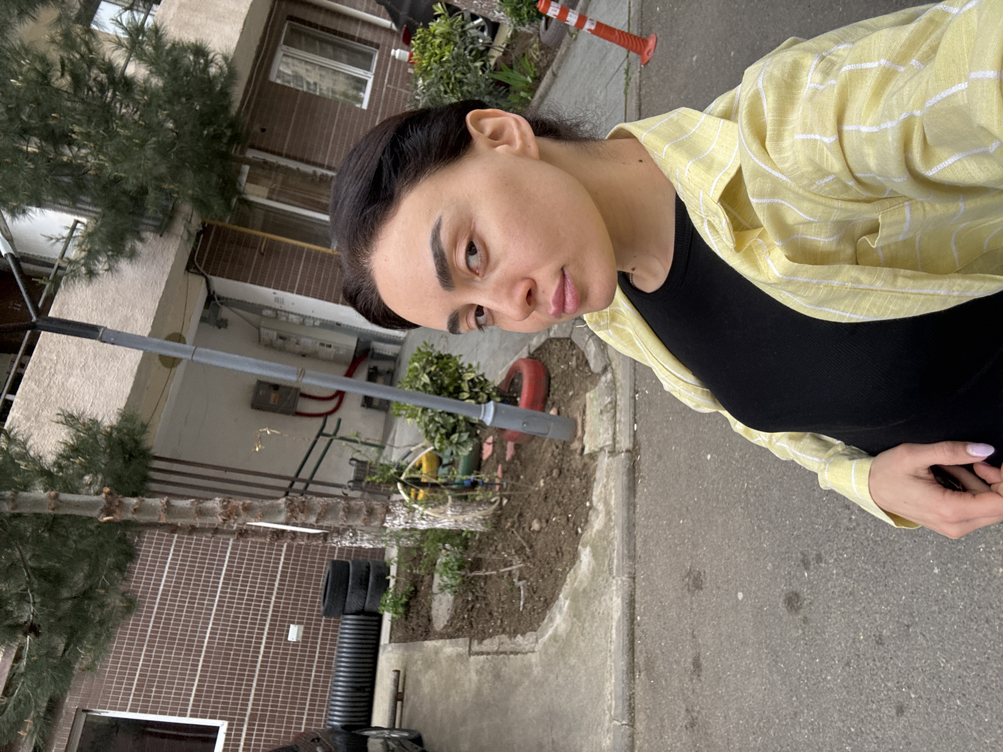 A daytime outdoor portrait features a woman standing on a paved surface in an urban setting, likely in Tbilisi, Georgia. She is positioned towards the right side of the frame, looking directly at the camera with a neutral expression. Her dark hair is pulled back, revealing her ears. She has dark eyebrows and natural-looking makeup. She wears a black ribbed top under a light yellow button-up shirt with thin white stripes. Her left hand, showing light-colored manicured nails, rests in front of her.
The background consists of a multi-story building with a lower section covered in brown square tiles and an upper section of a lighter, possibly cream or beige, color. Windows are visible on the building. A prominent evergreen tree, possibly a pine, stands to the left. A tall, slender gray pole, possibly a street lamp or utility pole, extends vertically through the mid-ground. Near its base, a small patch of garden or unpaved ground contains some greenery and planter pots, including a red one. Other objects in the background include a black corrugated pipe, what appears to be stacked black tires, and a red and white striped traffic cone further in the distance. The ground is a mix of asphalt or concrete pavement and some dirt/vegetation. The lighting is soft and even, suggesting an overcast day or indirect sunlight. No visible text is present.