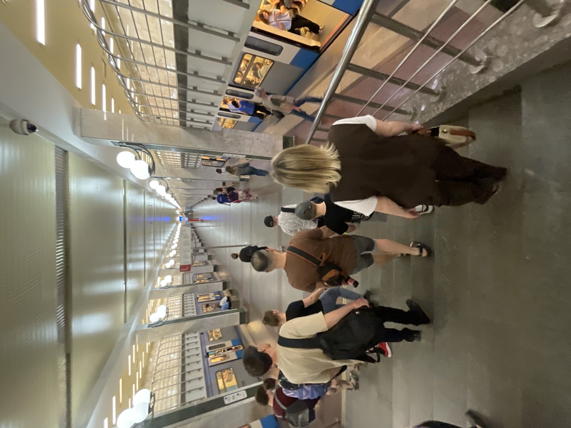 The image depicts a long, subterranean metro station platform in Moscow, Russia, viewed from an elevated staircase. Numerous individuals are present, engaging in various actions typical of a transit environment.
In the foreground, a group of people descends a wide, dark grey staircase, which has a metal handrail on the right side. The closest person to the camera, walking down the stairs with their back turned, is a woman with blonde hair, wearing a dark brown vest over a white shirt and dark pants, holding a light-colored handbag with a brown strap in her right hand. Behind her, other individuals, predominantly male and casually dressed, also descend the stairs, some carrying backpacks.
The station platform extends deep into the background, flanked by light-colored walls and numerous pillars. Along the length of the platform, several blue and grey metro trains are stopped, with their doors open, indicating boarding and alighting of passengers. People are visible entering and exiting the train cars and standing on the platform.
The station's ceiling is high and features multiple lighting types: large, white spherical globe lights are mounted on the walls and pillars, and recessed strip lighting runs horizontally along the ceiling. Above the track level, a distinctive metallic grate or lattice-work structure runs along the length of the platform. A dome-shaped security camera is affixed to the ceiling directly above the staircase in the upper left of the frame. The overall environment appears well-lit and maintained.