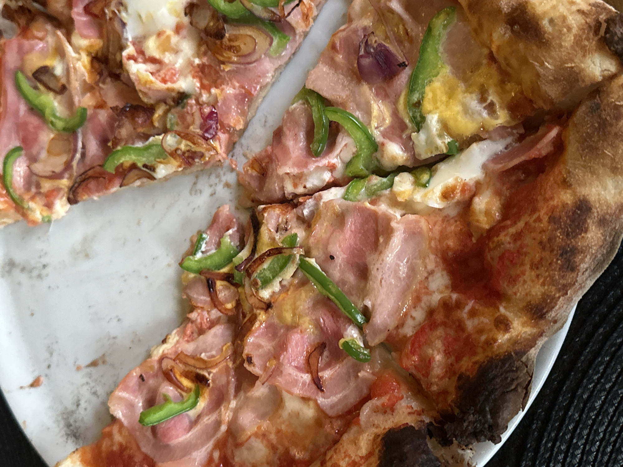 The image shows a partially consumed pizza on a white plate. The pizza has a thick, charred crust. The toppings include ham, tomato sauce, mozzarella cheese, green bell peppers, and caramelized onions. The background appears to be a dark surface, possibly a table. There is no evidence in the image to suggest that this is in Sofia, Bulgaria.
