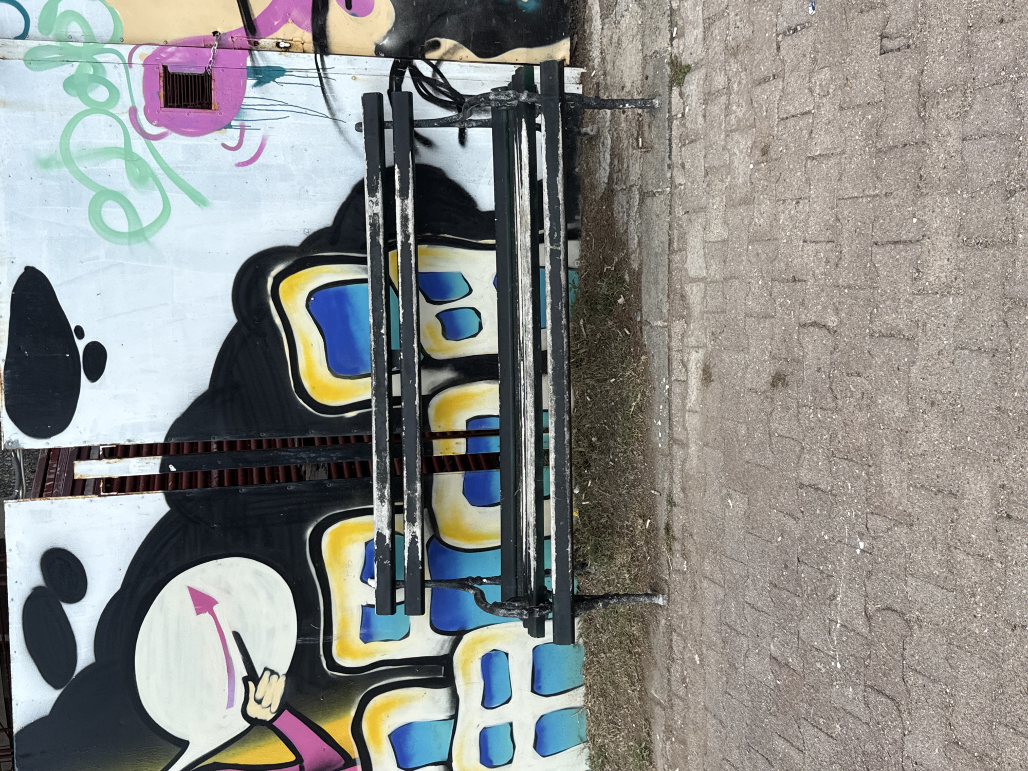 The image shows a section of graffiti art on a wall next to a cobblestone pathway. The graffiti features bold colors, including black, white, yellow, blue, and pink, with abstract shapes and what appear to be stylized windows or eyes. One section of the graffiti depicts a white circle with a pink arrow pointing upwards and a hand reaching towards it, possibly representing progress or a goal. A metal grate or fence is positioned in front of some of the graffiti. The setting appears to be outdoors, with the cobblestone path suggesting a public area or walkway. The lighting indicates it is daytime, and the weather seems fair. The overall mood is vibrant and urban, typical of street art. No discernible text is visible in the image.