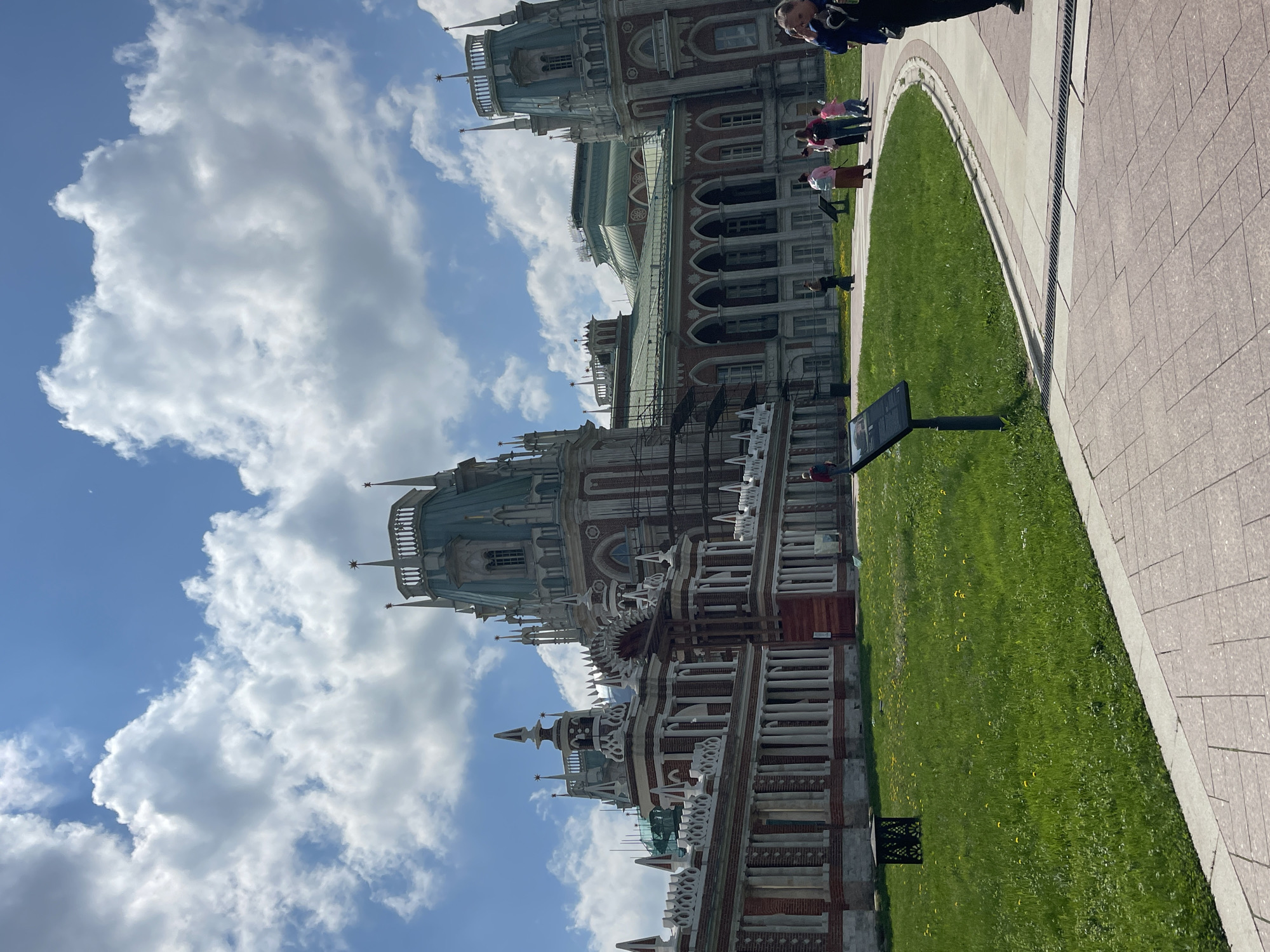 The image captures a view of the Tsaritsyno Palace in Moscow, Russia. The palace exhibits ornate architecture with red brick walls, white decorative elements, and multiple spires. To the right of the palace, there is a grassy area with a paved walkway running alongside it. Some people are walking along the path. The sky is blue with scattered white clouds. A black sign is visible on the grass near the path.