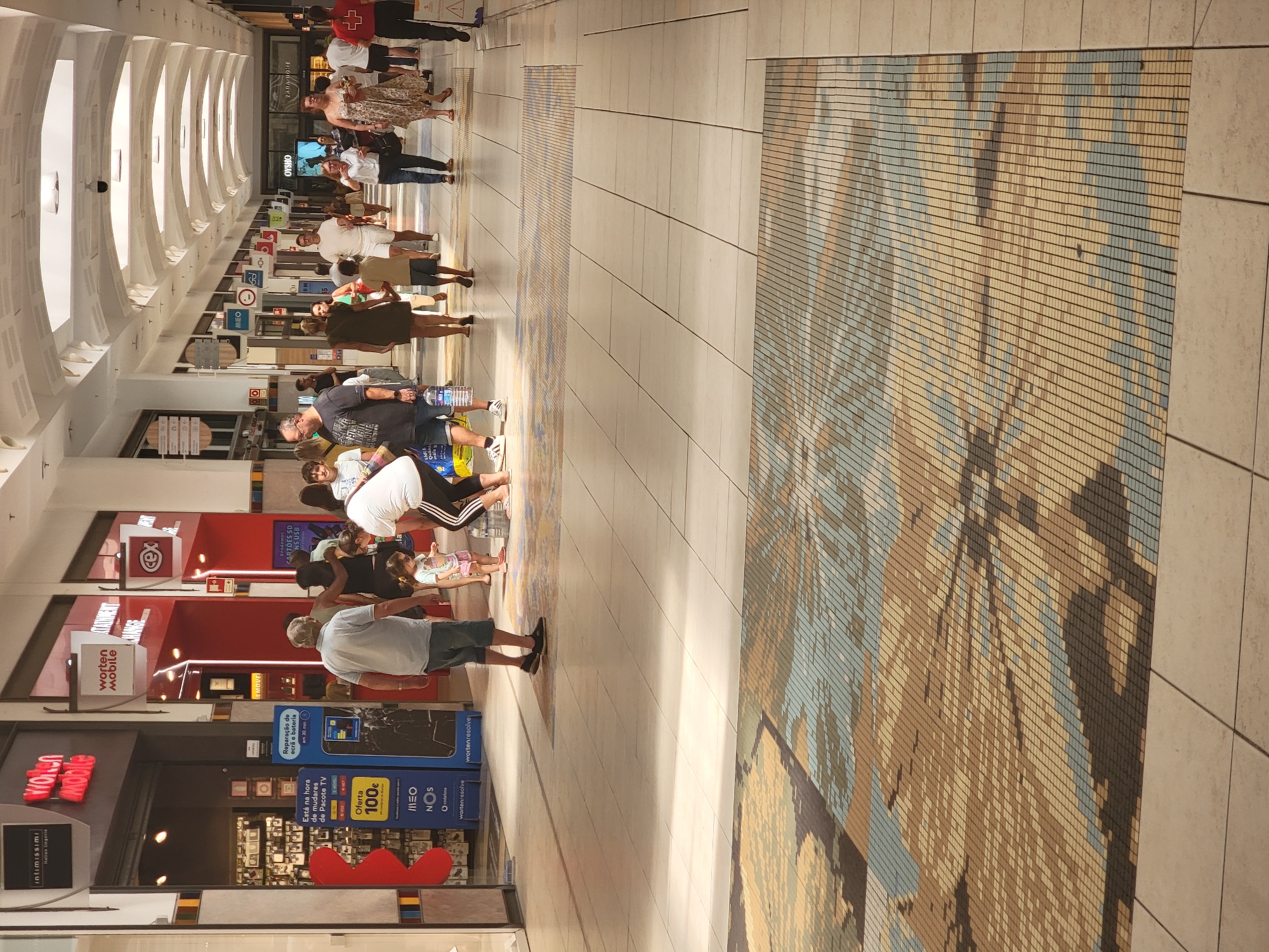 The image shows the interior of a shopping mall in Faro, Portugal, specifically the Forum Algarve mall. The scene captures a portion of a wide, brightly lit corridor with a patterned tiled floor. In the foreground, a large section of the floor features a mosaic design in shades of brown, beige, and light blue, creating an abstract pattern. Several people are visible walking and interacting within the mall.
On the left side of the image, there is an entrance to a store with signs for "Worten Mobile" and "Intimissimi Italian Lingerie." A blue promotional stand is visible, displaying information about phone repairs and offering a 100€ discount. Near this store entrance, a man wearing a light blue shirt and blue shorts stands facing away from the camera, looking towards a young girl in a pink dress who is also facing away. A woman in a white shirt and black pants with white stripes is partially visible behind the girl. Further down the corridor, various other shoppers are seen.
To the right of the main group, a man in a grey t-shirt and shorts walks away from the camera. In the background, more people are dispersed throughout the mall. Storefronts with illuminated signs are visible along both sides of the corridor, including "OYSHO" and "ZARA HOME." The ceiling of the mall is arched and has multiple light fixtures. The overall atmosphere appears to be that of a typical, busy shopping environment.