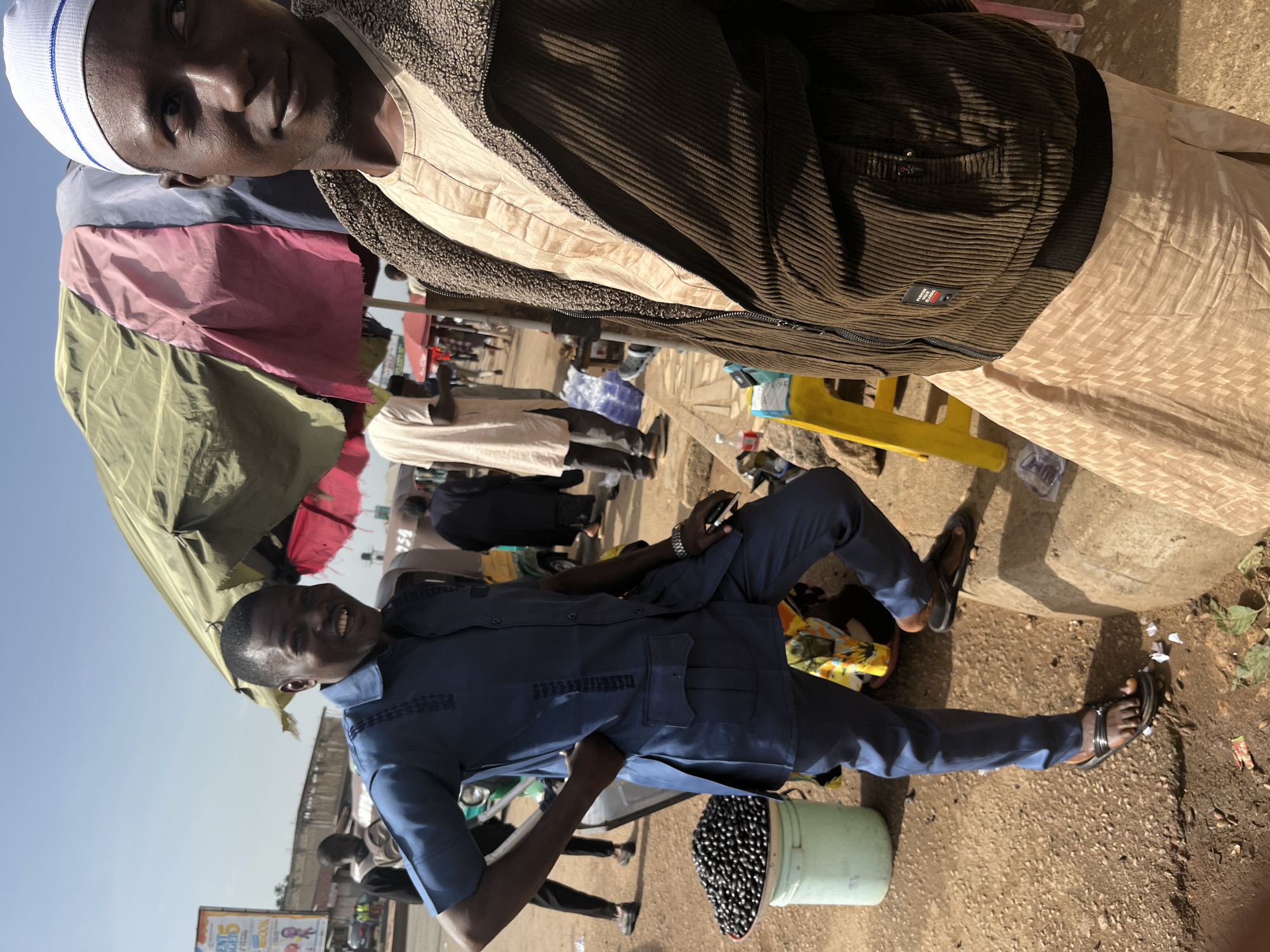 In Gura Top, Nigeria, two men are visible in a bustling outdoor market. The man on the left, wearing a dark blue traditional African outfit and sandals, is smiling broadly with his hand on his hip. Beside him, a bucket filled with dark, round objects, possibly fruit or beads, is present. The man on the right, facing away from the camera and wearing a white cap, a beige garment, and a brown corduroy jacket, is captured in a profile view. The background reveals a street scene with more people, vehicles, and market stalls covered by colorful tarpaulins. The ground is dusty and uneven, characteristic of a lively marketplace.