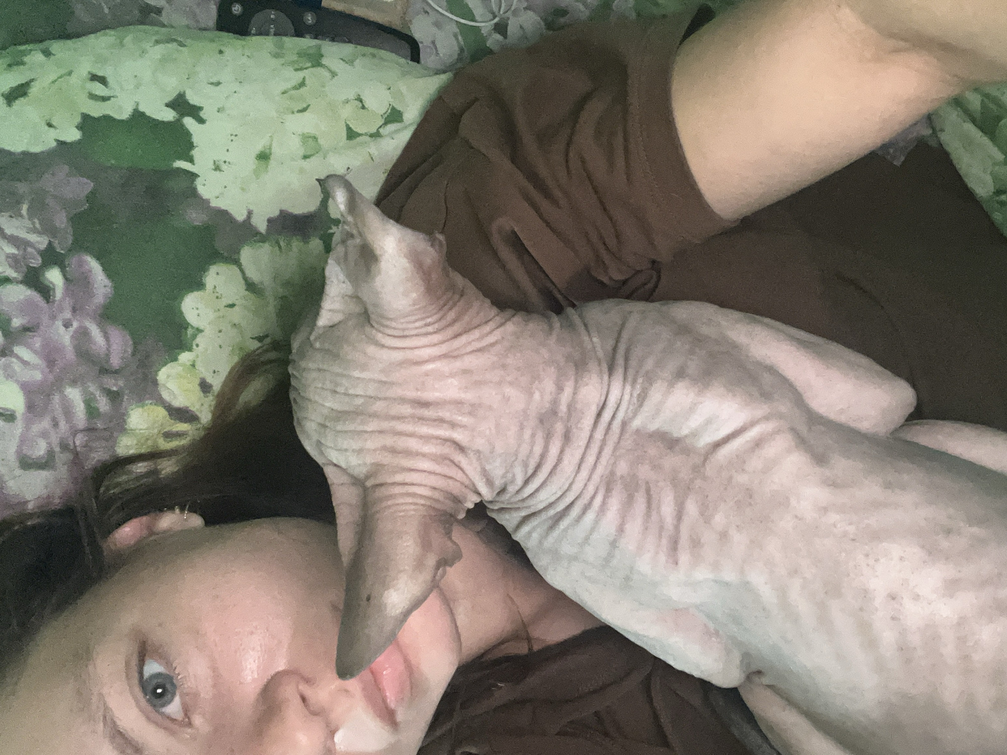 A woman and a hairless cat are visible in a selfie. The woman has fair skin and an eye is visible; it appears blue. A hairless cat is lying on or beside her, its head near her face. The woman is wearing a brown top. They are lying on a floral patterned bedspread, and an electronic device is visible in the background. No information about the location in Nizhny Tagil, Russia, can be derived from the image.