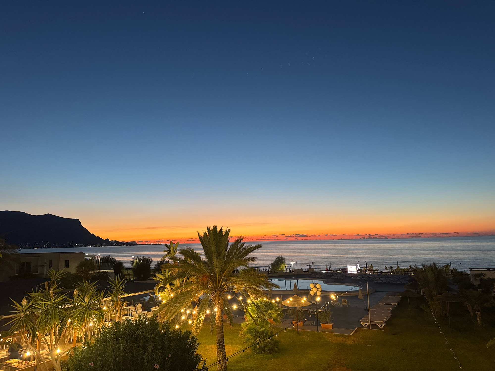 This is a high-angle, wide shot of a coastal resort area at dusk. The sky transitions from a deep blue at the top to a vibrant orange and yellow at the horizon, indicating sunset. The sea is visible in the background, meeting the colorful sky. In the mid-ground, a swimming pool area with lounge chairs and umbrellas is illuminated by artificial lights. Several palm trees and other tropical vegetation are scattered throughout the resort grounds, some decorated with string lights. A dark, mountainous landmass is visible in the far left background. The scene appears to be set in the town of Isola delle Femmine, Italy, likely a resort or hotel overlooking the coast. There are no people clearly visible in the image.