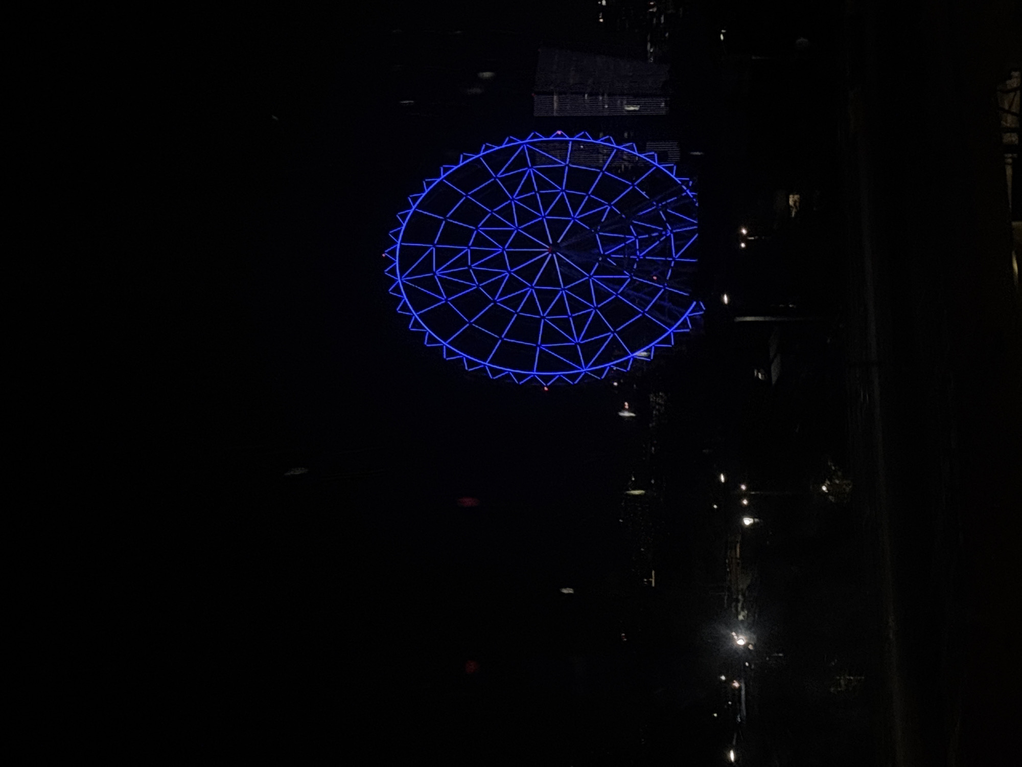 The image shows a partially visible Ferris wheel illuminated in blue against the dark night sky. It is located in Mexico City, Mexico. There are dark, indiscernible buildings behind the Ferris wheel, and faint lights can be seen along a street.