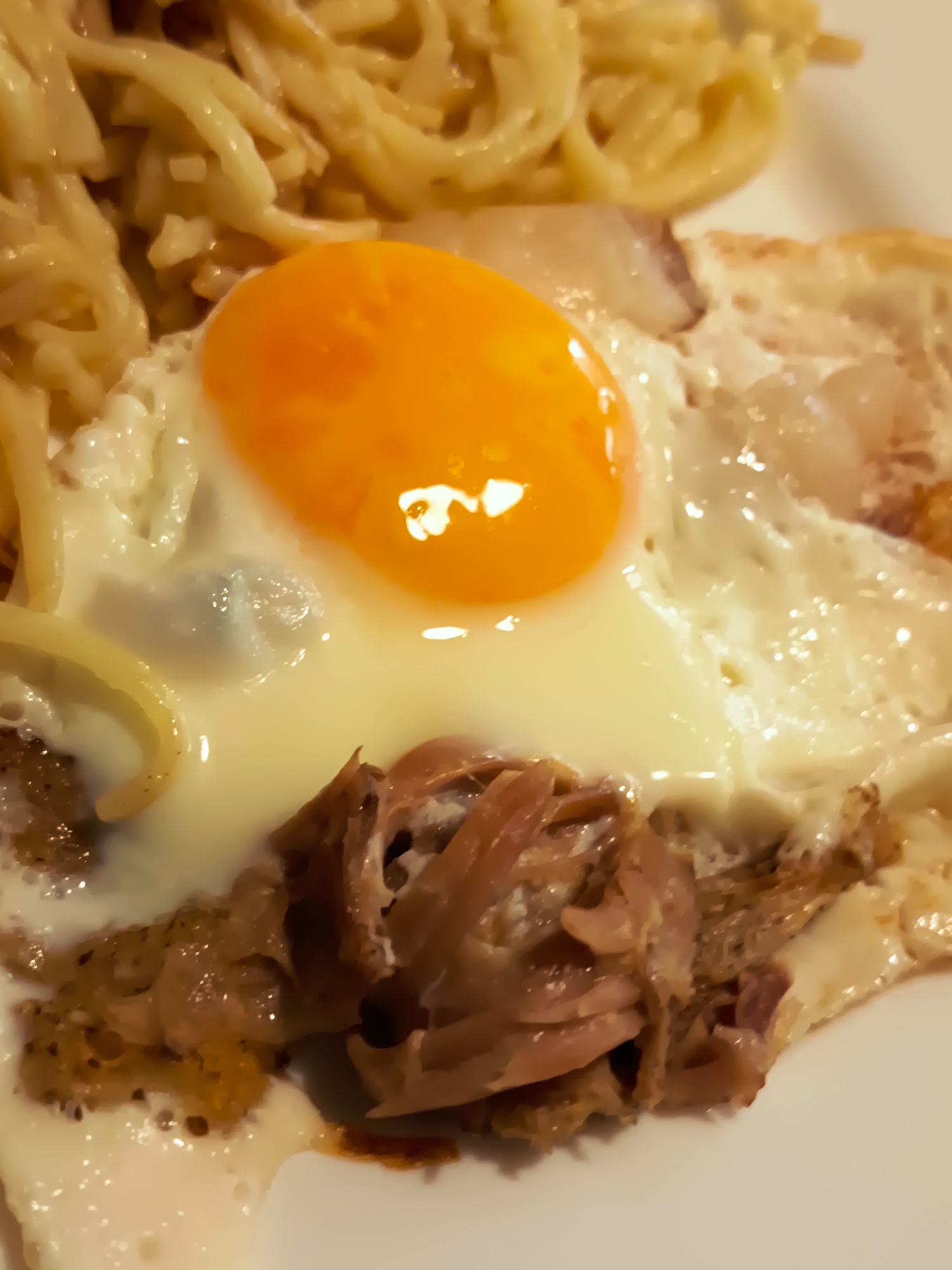 A close-up, eye-level, medium shot shows a plate of food, with noodles, a fried egg, and shredded meat. The egg is sunny-side up, with a bright orange yolk and cooked white. The noodles appear to be spaghetti, and the meat is brown and shredded. The food is on a white plate, and the background is out of focus. The lighting is bright, and the colors are vibrant. There is no indication of the city of Izmail, Ukraine in this image.