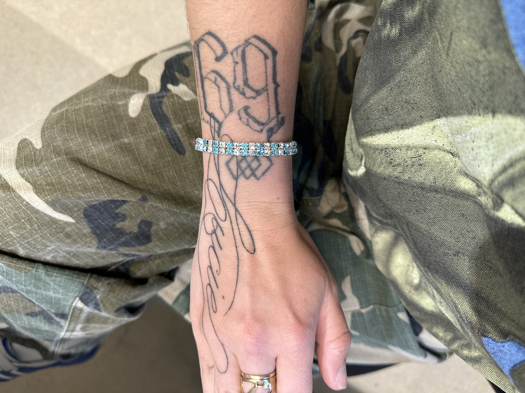 The image shows a person's left arm and hand resting on their lap. The person is wearing camouflage patterned pants. The arm has two prominent tattoos: a large "69" on the forearm and a cursive inscription on the hand. A two-row bracelet with clear and blue stones adorns the wrist. A gold ring is on the ring finger. The background is out of focus, but hints at a light, possibly neutral-colored interior. There's no evidence to suggest that this image was taken in Paris, France.
