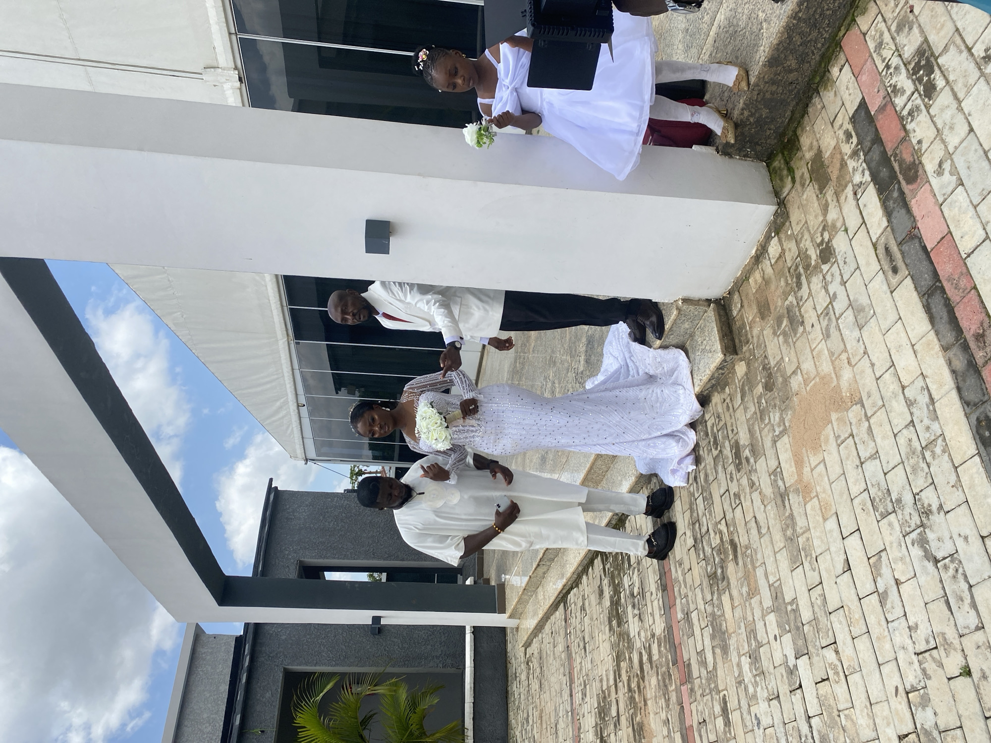 A wedding party is posing for a photograph outdoors, near a modern building with large glass windows and a white awning. The bride, wearing a sequined white gown, stands with a bouquet of white flowers. Beside her is the groom, dressed in a white traditional Nigerian outfit. To their left, another man in a white traditional outfit holds a phone. To the right, a young girl in a white dress, holding a small bouquet, stands with her back to the camera. The ground is paved with interlocking bricks, and there are steps leading up to the building. The sky is partly cloudy.