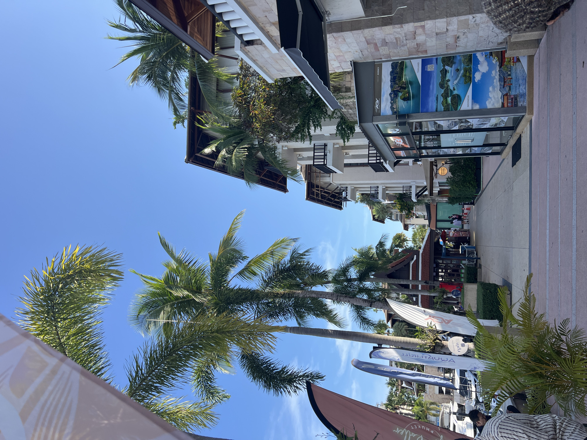 The image depicts a street scene in Ko Kaeo, Thailand, with a clear blue sky. Tall palm trees line the walkway, with their fronds extending towards the sky. On the right side of the image, a multi-story building with white walls and dark balconies is visible, adorned with light-colored stone accents. Below the building, a storefront displays travel posters showcasing scenic island destinations, identified as Thailand and Indonesia. A black awning extends over part of the storefront. To the left, a series of tall, slender flags, some bearing logos and text, stand in front of a row of boats docked in a marina. Several people are walking along the paved pathway, and a man in a striped shirt is seated on the left side of the frame. The environment appears to be a resort or a harbor area.