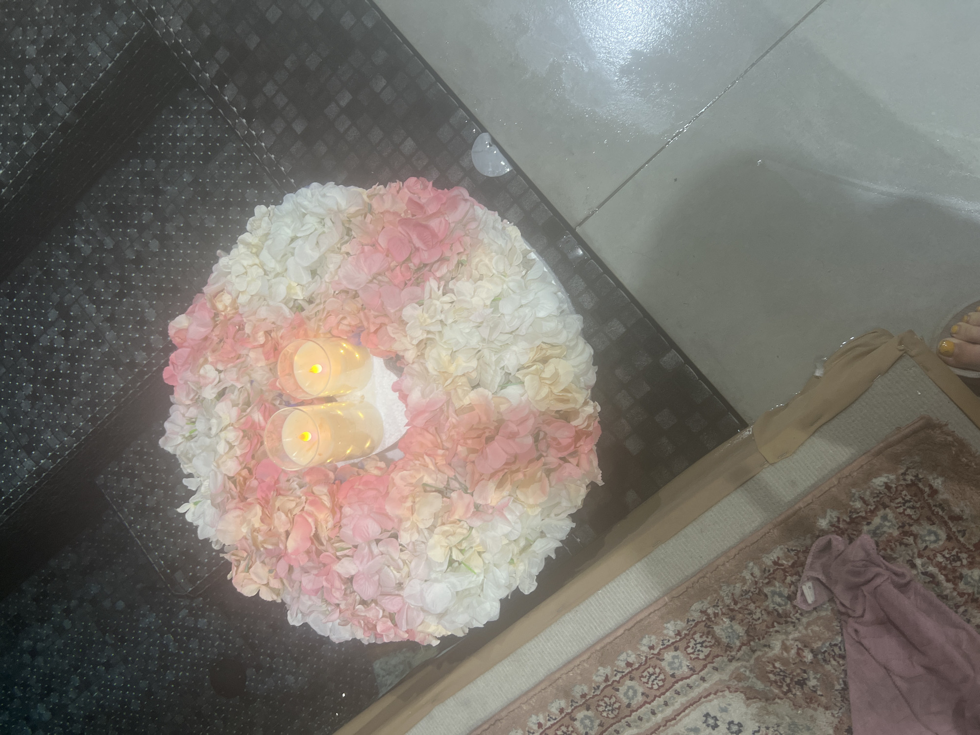 In the city of Hof HaCarmel Regional Council, Israel, a circular floral arrangement, composed of pink and white artificial flowers, is placed on a dark, textured surface, possibly a table. Two clear glass votives, each containing a lit candle with a yellow flame, are nestled within the arrangement. To the left of the arrangement, a portion of a rug with a floral pattern in muted pinks, reds, and blues is visible. A pink garment is draped on the rug. The background shows a tiled floor, some of which appears wet.