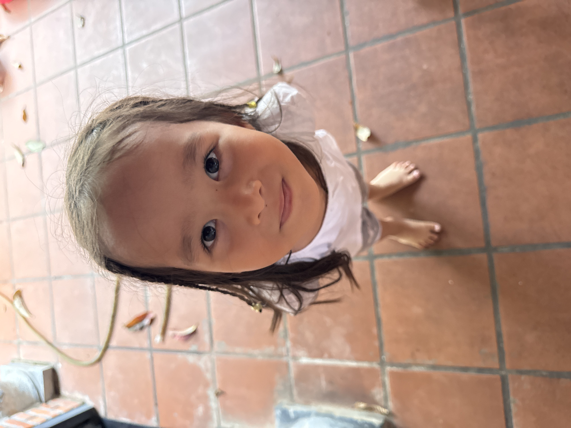 A child with dark hair and light skin looks up at the camera from a low vantage point. The child stands on a terracotta-tiled floor. The tiles are large, rectangular, and separated by dark grout lines. Some scattered leaves are visible on the floor, and there appears to be an object partially visible in the bottom left corner of the frame that includes a green hose. The location is most likely Mũi Né, Vietnam, as it was specified in the prompt.