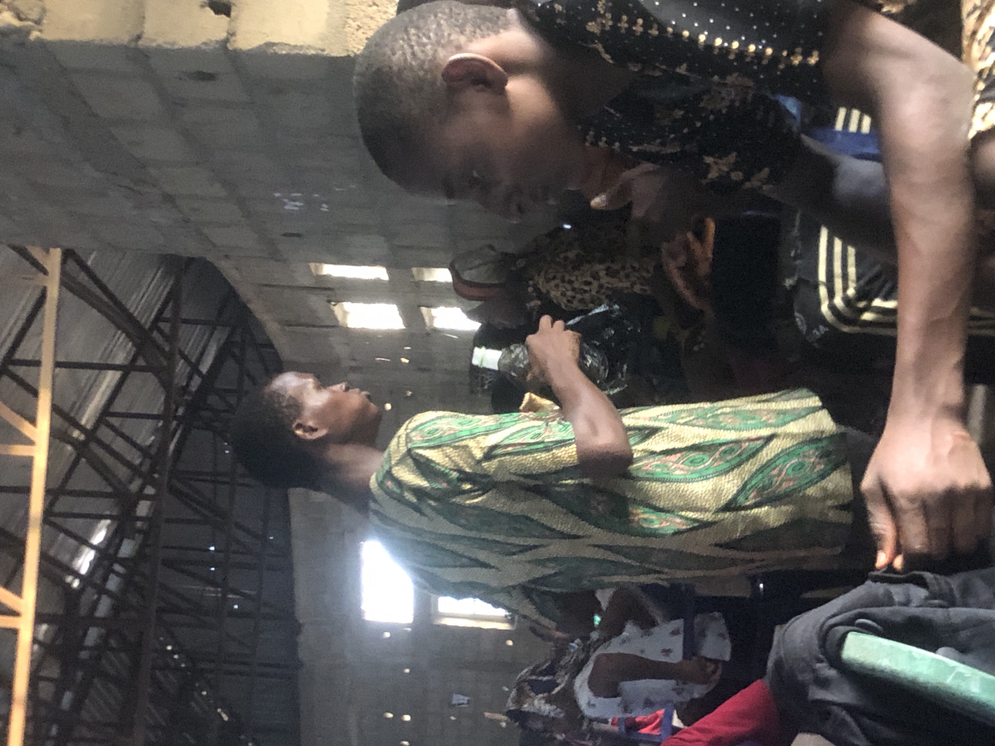 Two dark-skinned individuals are in close proximity, one wearing a green and yellow patterned shirt and the other wearing a black shirt with small gold designs and Adidas pants. They are indoors, appearing to be in a structure with a metal frame and what seems to be a cement ceiling. Based on the location provided, this is in Nteje, Nigeria.