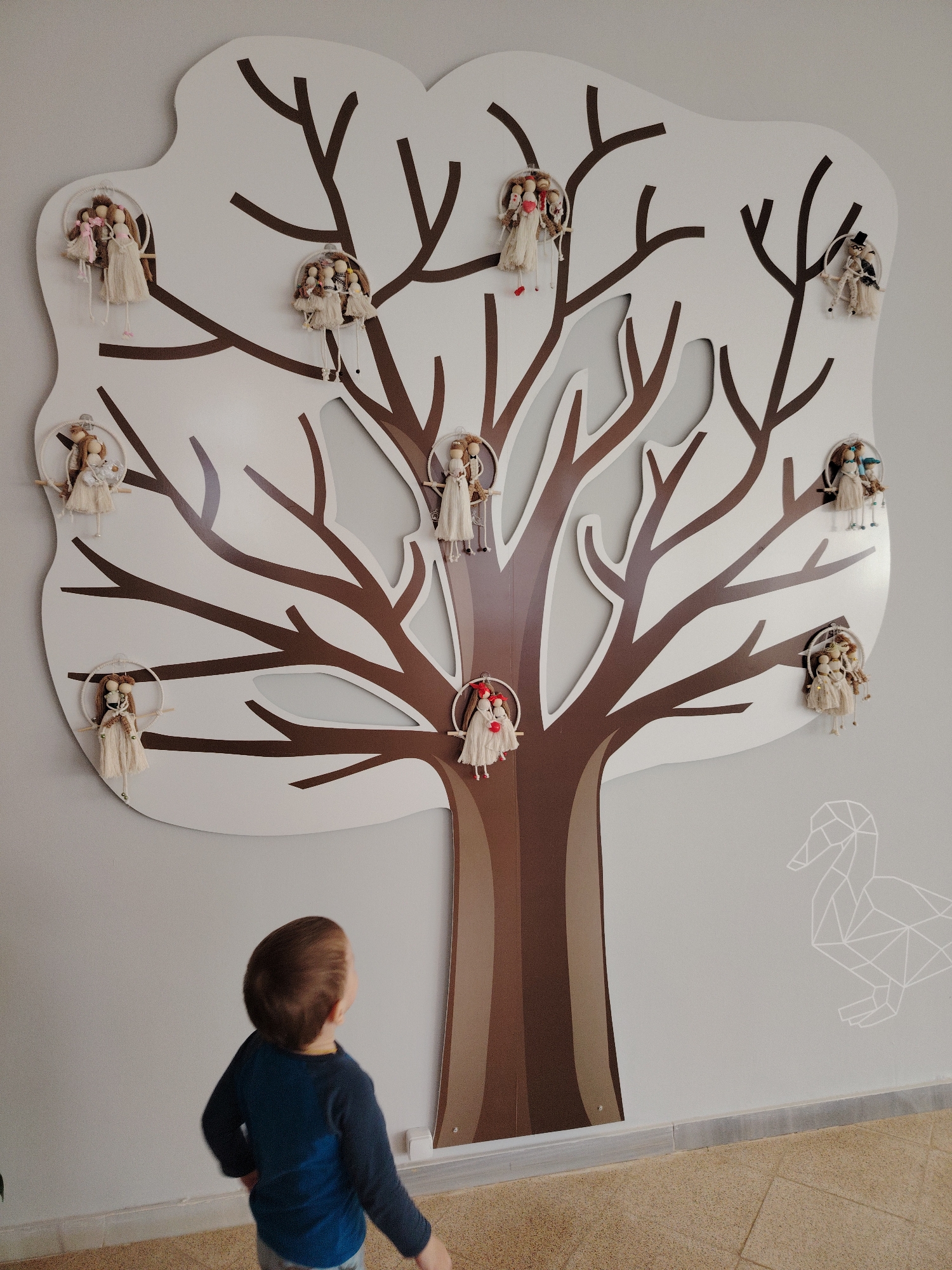 A small child stands in front of a wall decoration shaped like a tree in Markovskii, Russia. The tree is primarily white with brown branches and trunk. Hanging from the branches are numerous small dolls. To the right of the tree is a minimalistic geometric duck drawing on the wall. The child is facing the tree, wearing a blue shirt. The floor is tiled.