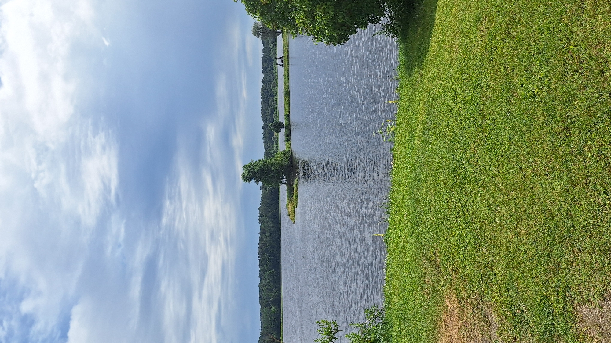 The image captures a serene waterscape in Stukmaņi, Latvia. A wide body of dark, rippling water dominates the center of the frame, bordered by lush green grass in the foreground. Across the water, a dense forest forms a dark green horizon line under a sky filled with dramatic clouds. A small, tree-covered island juts out into the water on the left side of the frame, with a solitary, tall tree standing prominently on it. Further in the distance, a swing set is visible on the opposite shore. The overall atmosphere is one of natural tranquility.