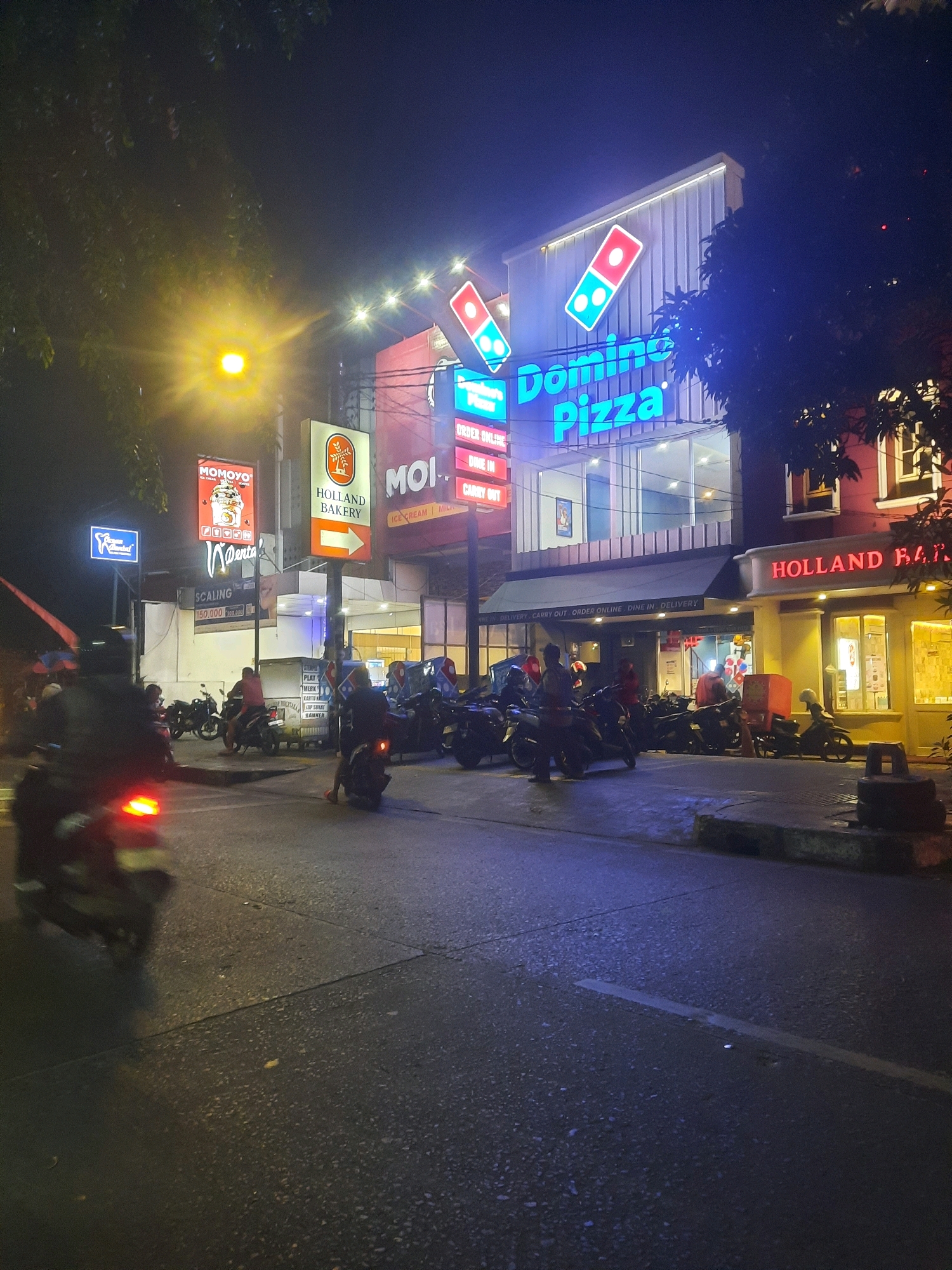 The image captures a nighttime street scene in North Jakarta, Indonesia, likely near the PIK 2 entertainment complex, characterized by vibrant neon signage and a bustling atmosphere. The focal point is a Domino's Pizza establishment, its bright blue and red logo prominently displayed on the building's facade. Adjacent to it, a "Holland Bakery" sign adds to the commercial landscape.
Several individuals are visible, predominantly men, engaged in various activities. Some are riding motorcycles, their headlights and taillights illuminating the dark street. Others appear to be standing or walking near the parked motorcycles, possibly waiting or socializing. One person is wearing a delivery box on their back, indicating food delivery services are active.
The environment is urban, with a paved road showing wet patches, suggesting recent rain. The street is lined with commercial buildings featuring large glass windows and illuminated interiors. Trees with dark foliage are present, partially obscuring the building lights and adding to the nighttime ambiance. The overall scene depicts a lively commercial area at night, with a focus on food establishments and transportation.