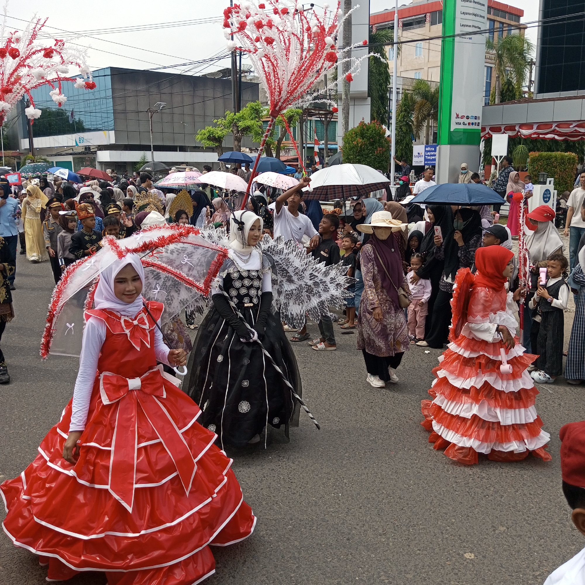 This is a ground-level, full-shot photograph of a parade taking place on a street in Lhokseumawe, Indonesia. The sky is overcast, and several people are holding umbrellas, indicating it may be raining or has recently rained. The parade features several participants in elaborate costumes, including two women in the foreground dressed as part of a procession.
The woman on the left wears a vibrant red dress with a large bow on the front, and a white long-sleeved shirt underneath. She is holding a clear umbrella decorated with red and white trim. To her right, another woman is dressed in a black gown with white floral patterns and accents, also holding a decorated umbrella. Behind them, a crowd of spectators, some holding umbrellas, watches the parade. Children and adults in various attire are visible, some dressed in formal or ceremonial clothing.
The background shows buildings, including a modern building with a glass facade and signage, and other structures that suggest an urban setting. A tall, vertical banner with green and white colors is prominent, featuring text in Indonesian. Trees and streetlights are also visible along the street. The overall scene depicts a festive community event, possibly a cultural or national celebration, given the colors and costumes.