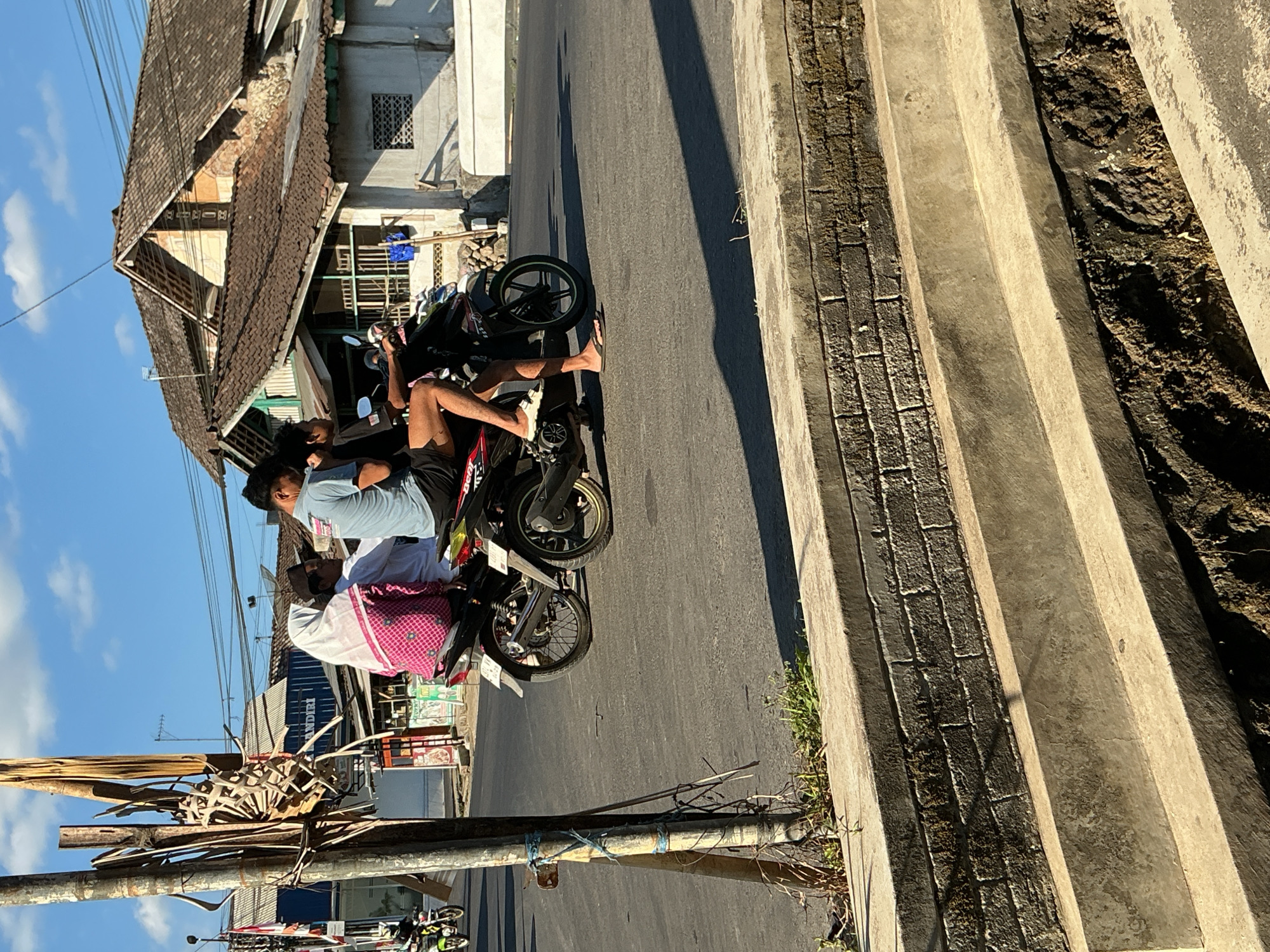 In Kebonrejo, Indonesia, on a clear, sunny day, three individuals are riding a single motorcycle on a paved road. The motorcycle is black and red, with two visible wheels. The rider in the front wears a light blue t-shirt and dark shorts. Behind him, a person in a white shirt and a dark cap is seated. The third person, in the back, is wearing a pink and white patterned headscarf and a pink garment, holding onto the rider in front.
The motorcycle is captured in motion, with the rider's legs visible, one wearing a sandal. The road is asphalt, with painted white lines. To the left of the motorcycle, a weathered wooden pole with dry, hanging vegetation is prominent, possibly a utility pole. In the background, traditional Indonesian buildings with tiled roofs are visible, along with power lines and streetlights. The foreground shows a concrete structure with brickwork below, possibly a drainage ditch or raised walkway, with some dry grass and dirt. The scene suggests a typical day in a residential or commercial area of the city.