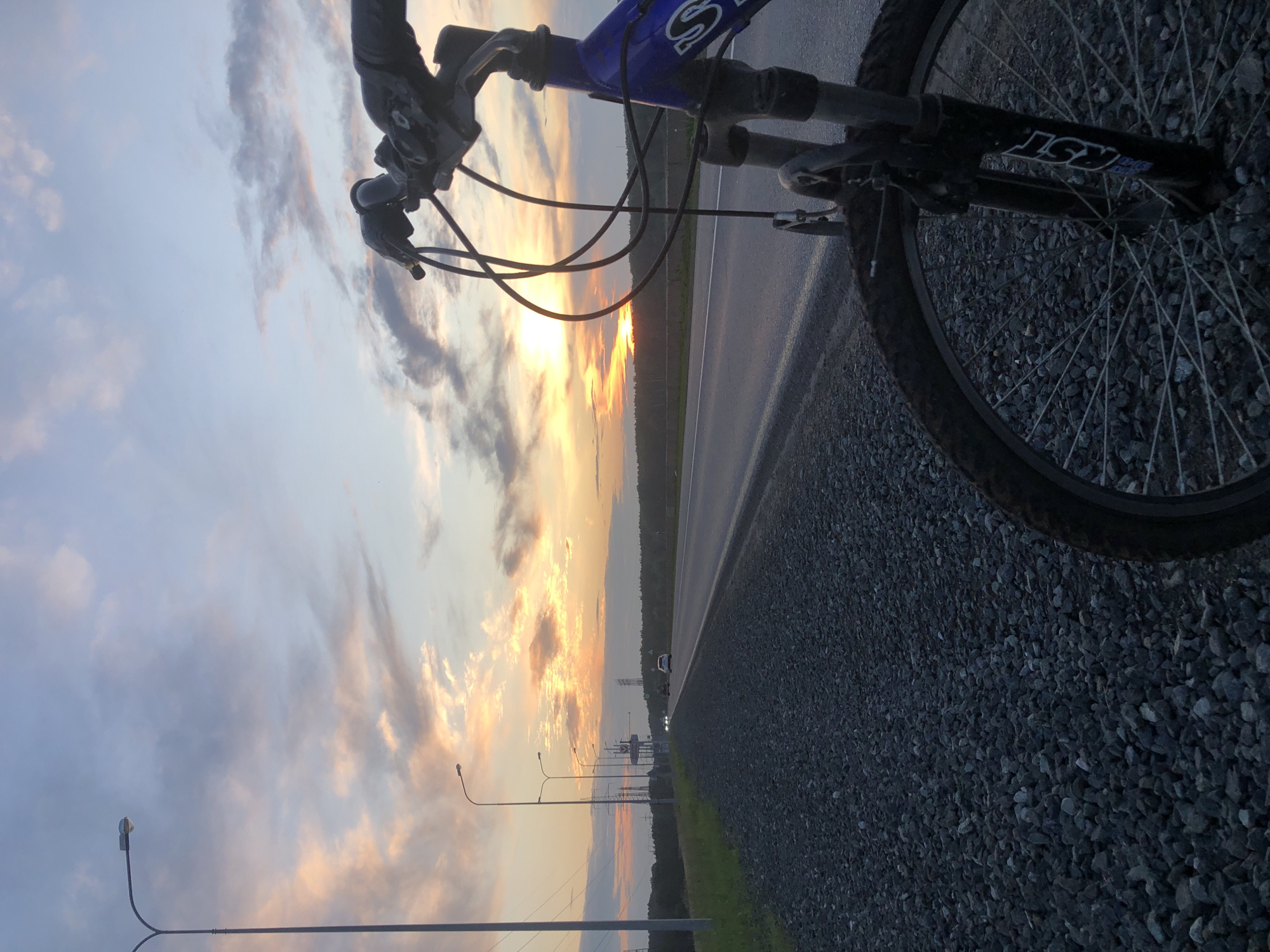 A bicycle is parked on the side of a highway in Chaikovsky, Russia, with the sunset visible in the background. The bicycle is blue and positioned in the foreground, with its handlebars and front wheel clearly visible. The highway stretches into the distance, with a few cars on it. The sky is filled with clouds, painted in hues of orange, pink, and purple by the setting sun. Streetlights line the side of the highway, and a forest can be seen in the distance. The ground is covered in gravel. The scene suggests a moment of pause during a bicycle ride at dusk.