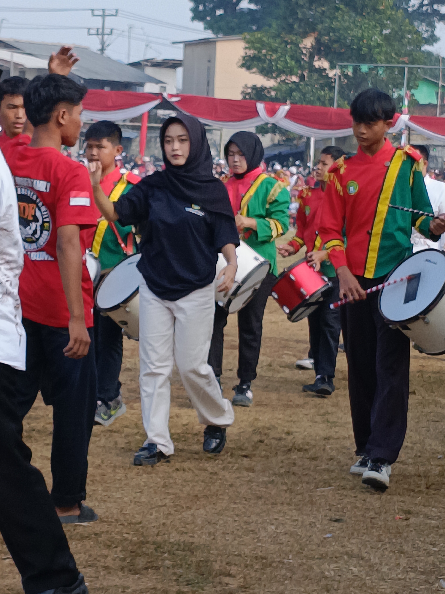 In Cibodas, Indonesia, a group of young people, likely in a marching band, are participating in an outdoor event. The foreground features several individuals, including a young woman in a black hijab and navy shirt, and a young man in a red shirt with a white and red Indonesian flag patch. To their right, two young men in red and green marching band uniforms are visible, one holding a drumstick and the other a red drum. Further back, other participants in similar uniforms can be seen. They are surrounded by spectators and decorations, including red and white banners. The ground is covered in dry grass and dirt, suggesting an open field or sports ground. The overall atmosphere appears to be one of a lively community gathering or performance.
