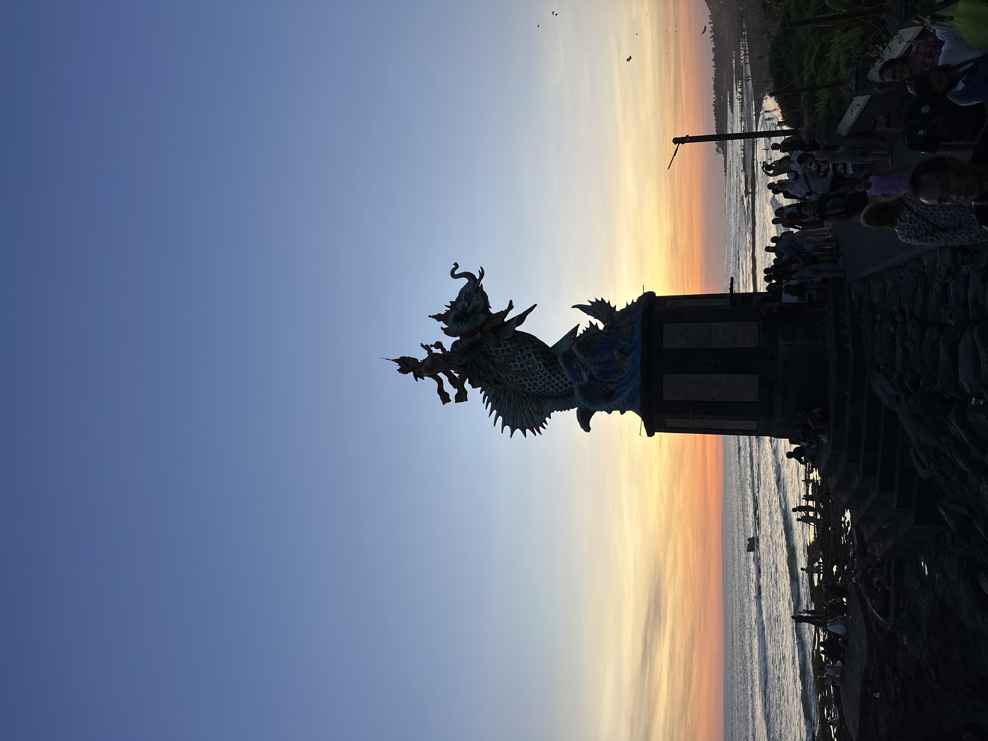 A statue sits near a group of people at a beach at sunset. The statue is of a dragon-like creature. The sky is a gradient of blue to orange and yellow. The people are standing on a stone walkway next to the beach. The waves are crashing in the background.