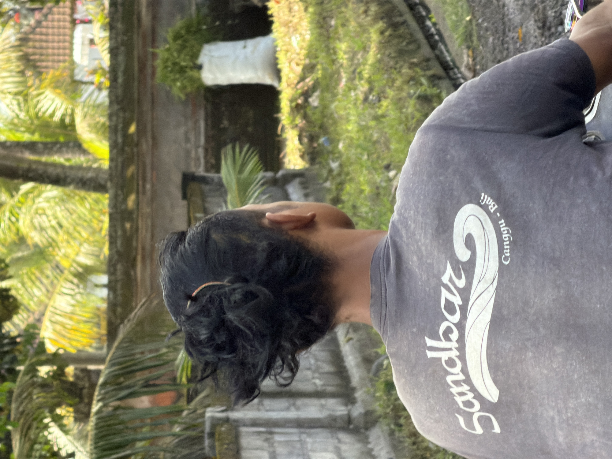 The image shows the back of a person wearing a gray t-shirt with a white graphic that reads "Sandbar Canggu - Bali". The person has dark, messy hair pulled back in a bun with a thin orange band. They are facing away from the camera, looking towards the right. The background appears to be an outdoor setting with lush green foliage, palm trees, and a weathered brick wall. In the distance, a white sack is visible among the greenery. The overall scene suggests a tropical or sub-tropical environment, consistent with the location in Mengwi, Indonesia. The lighting indicates it is daytime, possibly late afternoon due to the warm tones in the foliage.