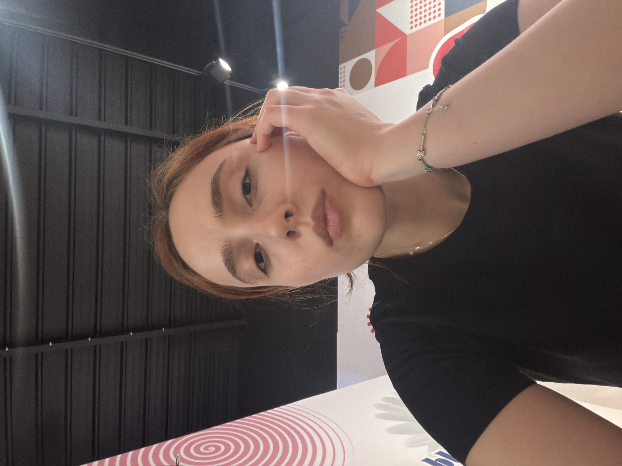 A young woman with auburn hair rests her chin on her hand, looking forward with a neutral expression. She is wearing a black t-shirt and a silver bracelet with small charms. The background features a dark, corrugated ceiling with lighting fixtures and a colorful patterned wall with geometric shapes and circular designs. The image appears to be a selfie taken indoors, possibly in a cafe or restaurant. There is no indication of the location being Strasbourg, France.