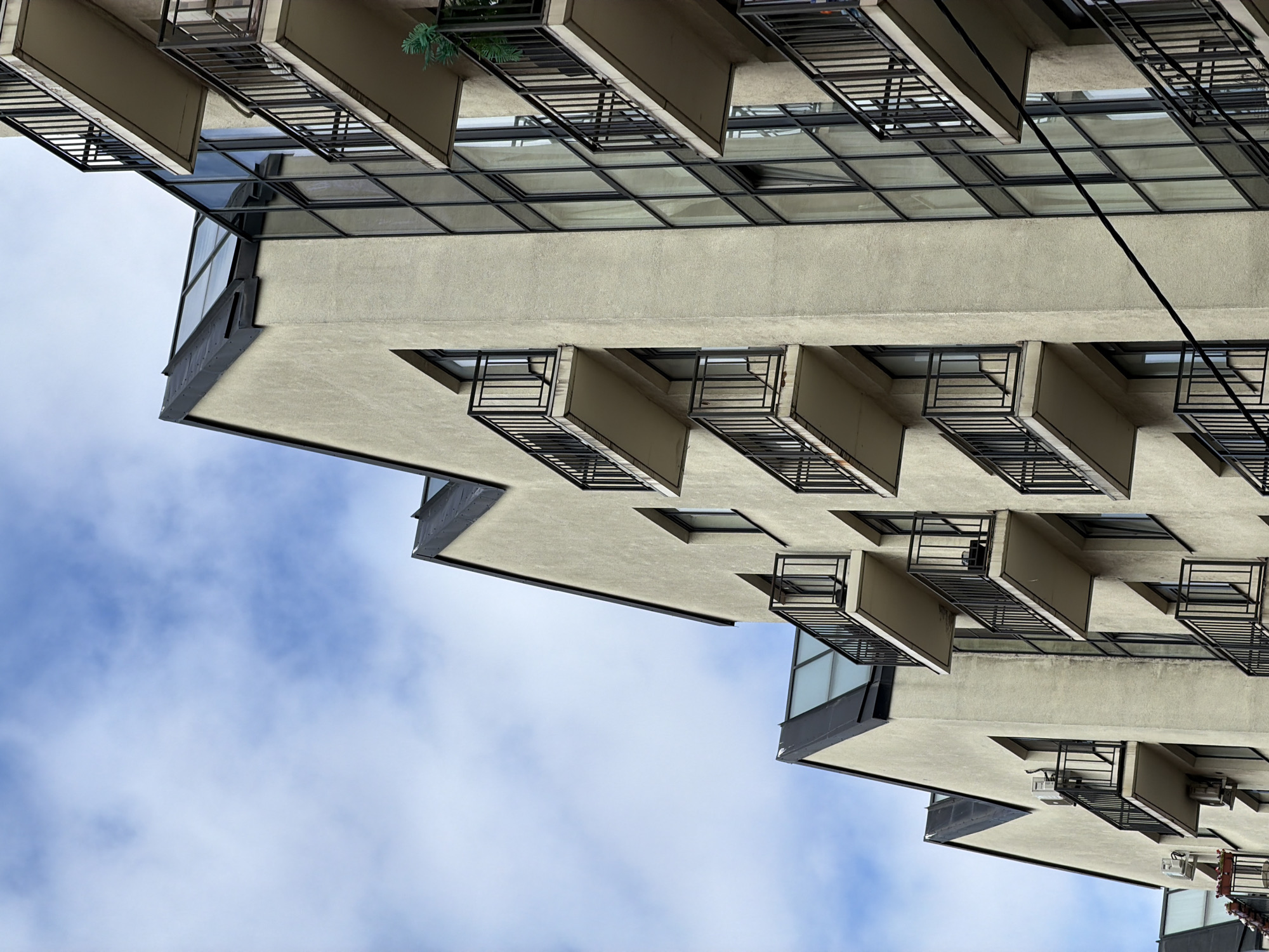 The image shows the exterior of a modern apartment building with a light beige stucco facade and dark-colored gabled roofs, some of which feature skylights. Several balconies with black metal railings extend from the building's facade. To the right, a section of the building features a large glass facade with dark mullions. The sky above is blue with scattered white clouds. The building appears to be located in Saint Petersburg, Russia, evidenced by the architectural style and the presence of an outdoor air conditioning unit mounted on one of the balconies. The scene is devoid of people or any overt activity, presenting a still, architectural view of the building against a cloudy sky.