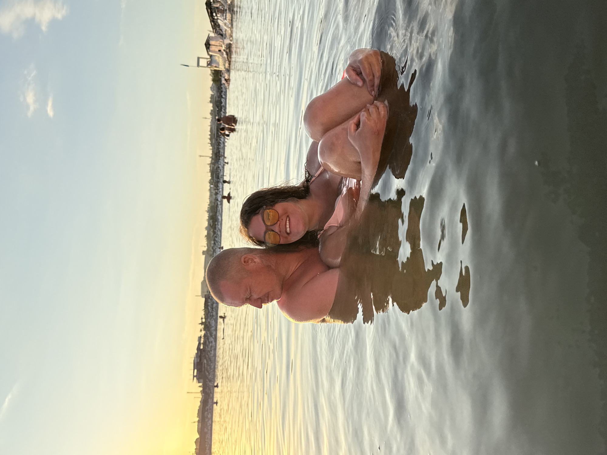 In the city of Sol-Iletsk, Russia, a man and a woman are in the water, with the man holding the woman. The man is on the left, looking to his right with his eyes closed and his mouth slightly open. He is shirtless and his head is bald. The woman is on the right, facing the camera with a smile and wearing sunglasses. She is holding her knees to her chest. They are both submerged in the water up to their chests. In the background, there are other people in the water and structures on the shoreline, including what appears to be a lifeguard tower and some buildings. The sky is a pale blue with a few scattered clouds, and the sun is setting on the left side of the image, casting a warm glow over the scene. The water is calm and reflects the sky and the people in it.