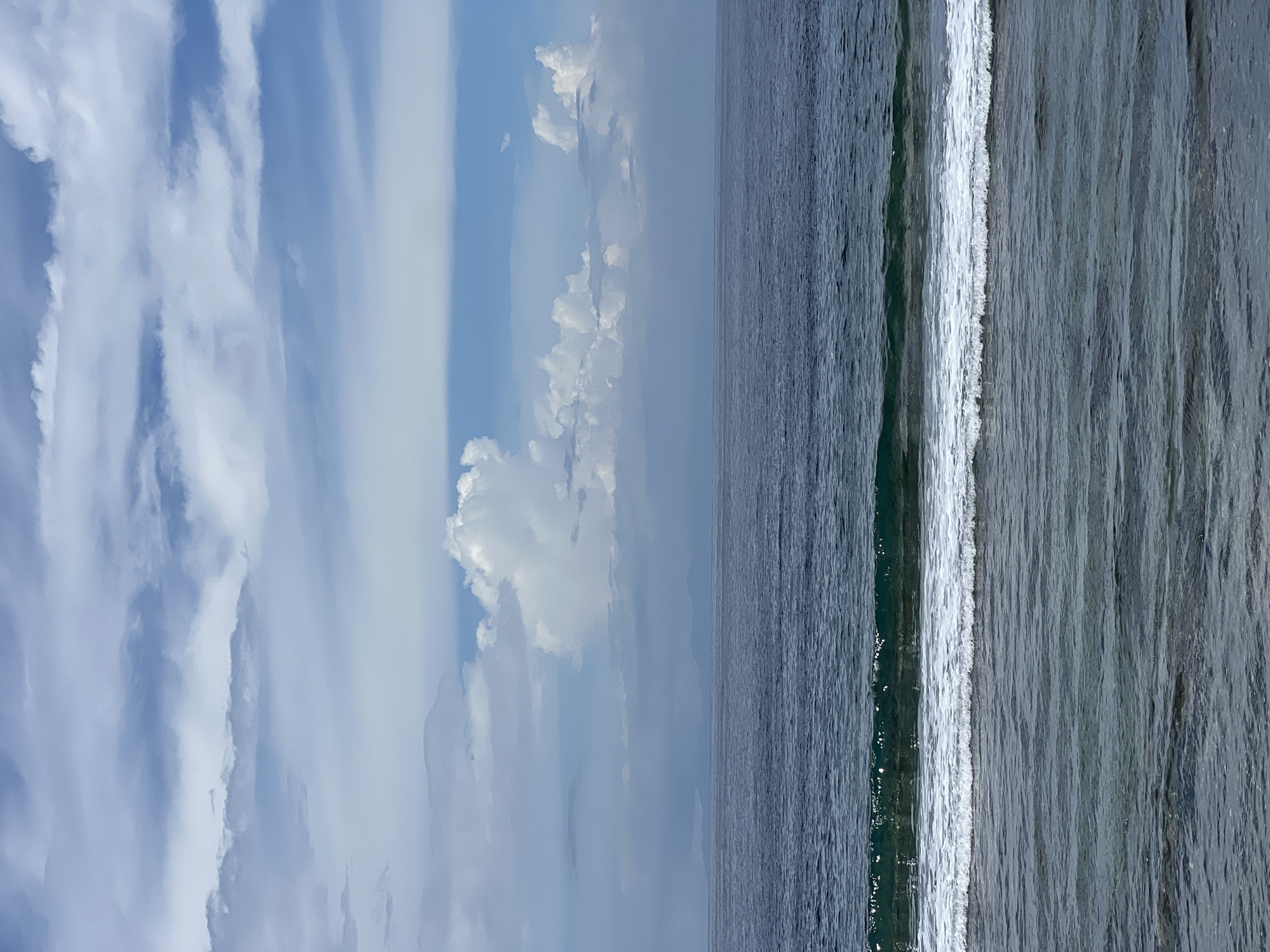 This media file presents a scenic landscape of the ocean and sky, likely captured in Nusa Penida, Indonesia. The upper portion of the image is dominated by a bright blue sky, adorned with a variety of white clouds. Some clouds appear wispy and elongated, while others are more defined and fluffy, suggesting a mix of cloud types.
The lower half of the image showcases the vast expanse of the ocean. The water is a deep blue-gray, with its surface textured by numerous ripples and small waves. A prominent white line of breaking surf stretches horizontally across the middle of the frame, marking a point where the ocean waves crest and foam. The water immediately surrounding this breaking wave appears a lighter shade compared to the deeper parts of the ocean.
The scene depicts a calm, clear daylight setting. No human activity, man-made objects, or visible text are present. The image conveys a sense of natural beauty and tranquility, focusing solely on the interplay between the sky, clouds, and the dynamic surface of the ocean.