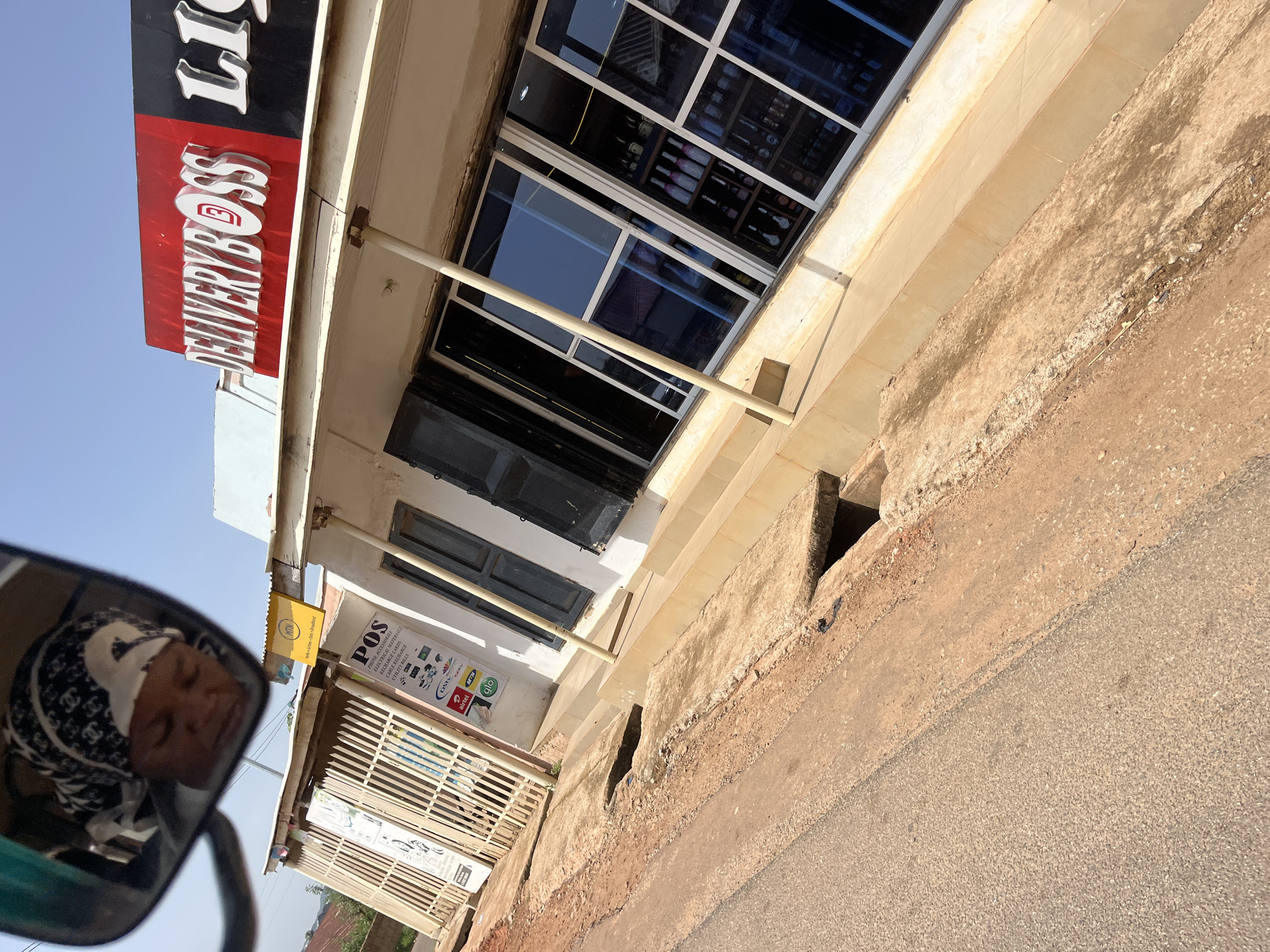 In Gura Top, Nigeria, a street-level view captures a storefront with a prominent red and black sign reading "DELIVERYBOSS LIQUOR." The store features large, reflective windows displaying shelves stocked with bottles of liquor, suggesting it is a retail establishment. A sidewalk with a drainage channel runs in front of the store. To the left, a metal gate partially obscures a business entrance. A yellow sign with the MTN logo is visible above the gate. In the reflection of a vehicle's side mirror, a person wearing a patterned headscarf is partially visible, their eyes closed, indicating they may be a passenger. The overall scene depicts a commercial area in Nigeria under a clear sky.