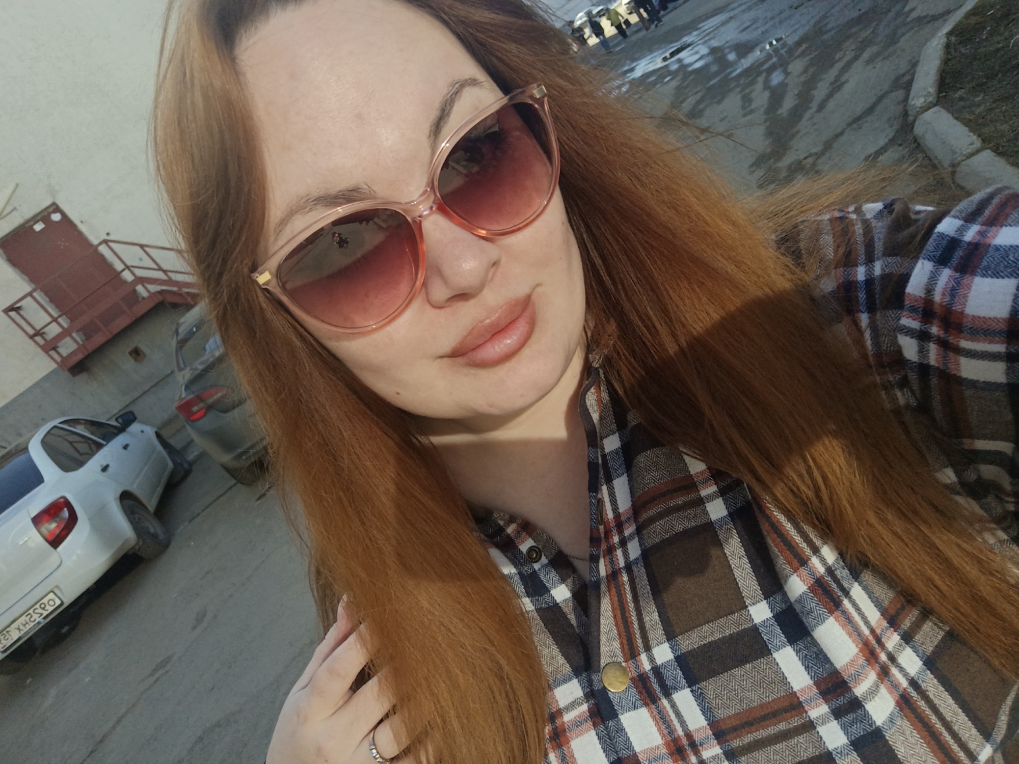 A selfie shows a woman with long, auburn hair and fair skin wearing pink-framed sunglasses and a brown, white, and red plaid shirt. She is smiling slightly at the camera.
In the background, there are parked cars and a building. A white car with the license plate "P882XH259O" is visible, indicating this image was taken in Russia. The location is likely the city of Chaikovsky, Russia, although the exact coordinates are not discernible from the image. The environment appears to be a paved area next to a building with a possible loading dock. The weather seems to be overcast.