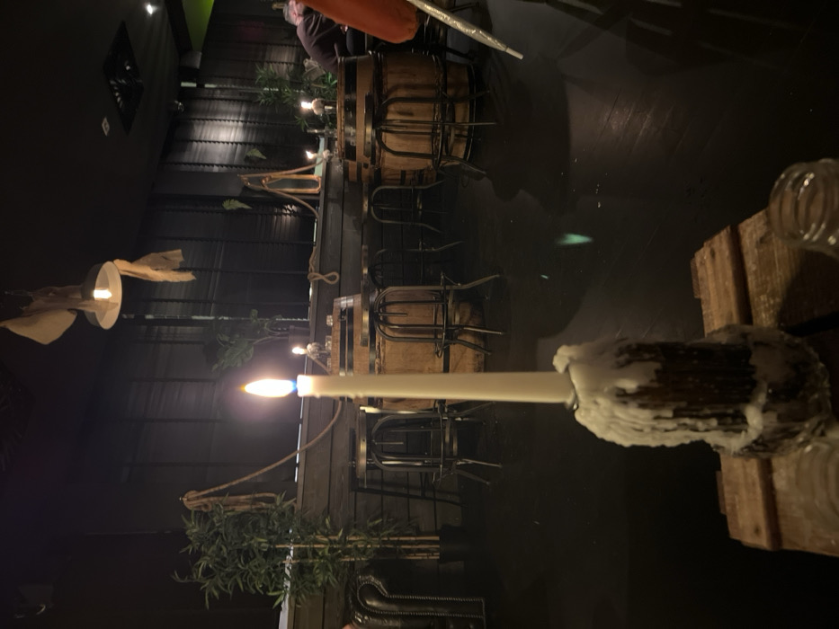 A dimly lit restaurant scene. A lit white candle is in the foreground, attached to a wooden stand. Several tables with wooden barrel bases and metal chairs are visible. The walls are dark, with textured, vertical panels. Sparse greenery decorates the tables. A dimly lit, propeller-like object hangs from the ceiling. There are other faint light sources.