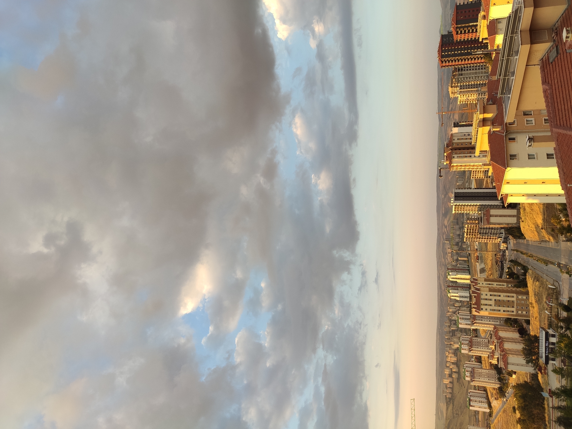 The image captures an aerial view of a city under a cloudy sky, likely Etimesgut, Turkey, during what appears to be the golden hour. The foreground is dominated by buildings with terracotta roofs. Further into the mid-ground, a dense cluster of apartment buildings, varying in height and color, lines the landscape. Some of these buildings are contemporary, with numerous windows and balconies, while others appear older. Construction cranes are visible amongst the buildings, suggesting ongoing development. A road winds through the urban area, with a few cars present. The background shows rolling hills and more distant residential areas extending towards the horizon. The sky is a dramatic mix of soft blue patches and large, diffused grey and white clouds, illuminated by the warm, soft light of the setting or rising sun. The overall impression is one of a growing urban environment bathed in the gentle light of dusk or dawn.