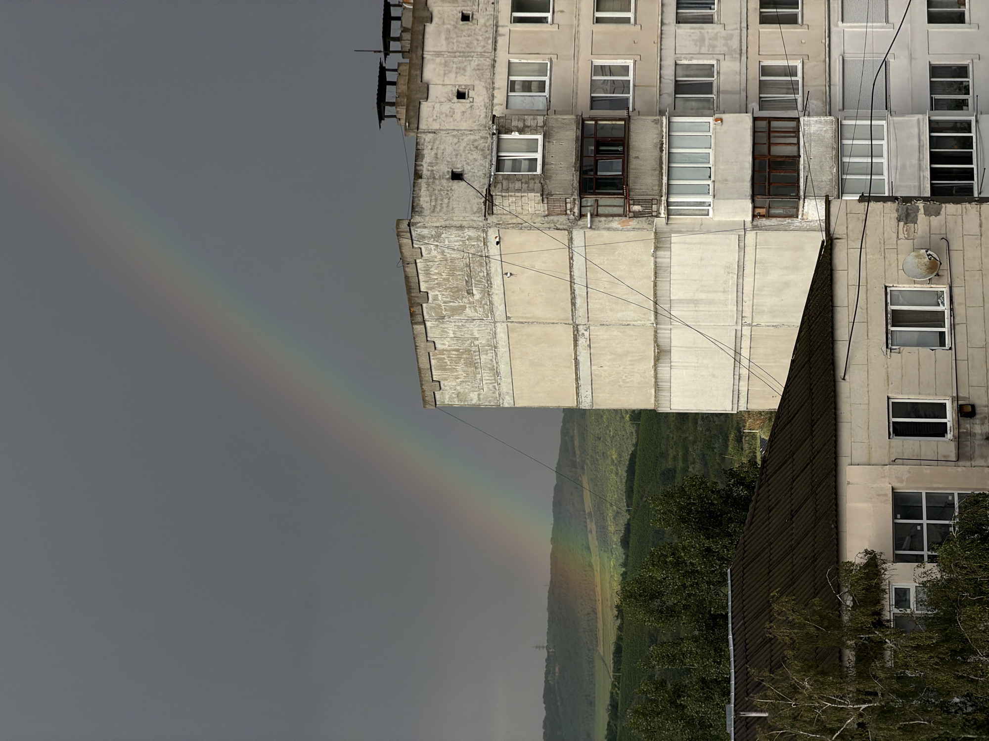 The image, taken in Strășeni, Moldova, depicts a view of residential buildings under a dramatic sky. A prominent rainbow arcs across the overcast sky, with its colors faintly visible against the dark clouds. In the foreground, a multi-story, concrete apartment building, showing signs of wear and tear, dominates the right side of the frame. Its facade is textured, with balconies featuring wooden-framed windows and metal railings. To the left and in front of the apartment building, a lower, dark-roofed structure is visible, with windows and a satellite dish attached to its side. In the background, rolling green hills, possibly agricultural land, are seen under the moody sky. The overall scene is one of urban residential architecture set against a natural, atmospheric backdrop.