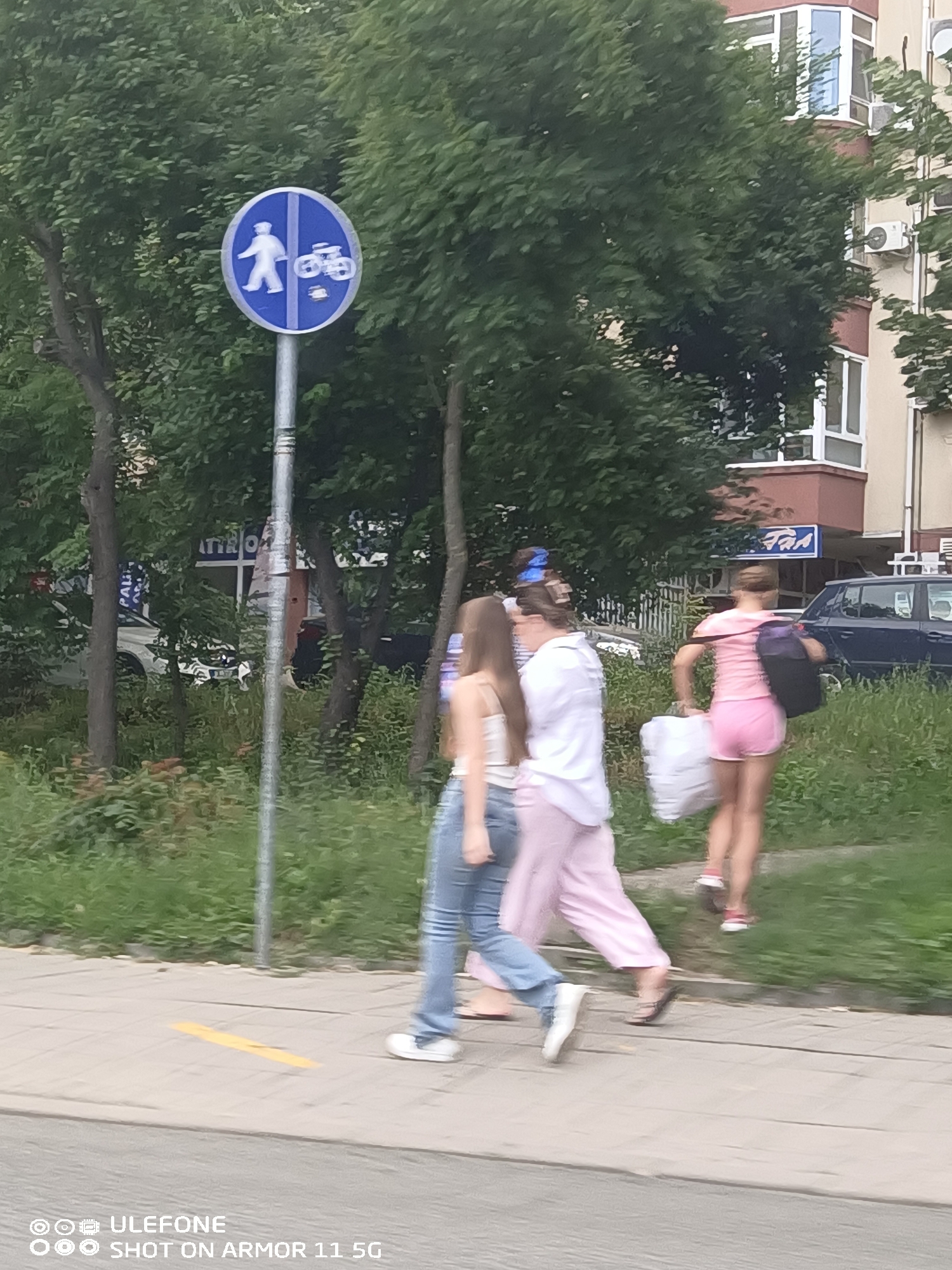 This image captures a daytime urban scene in an unknown city. In the foreground, a concrete sidewalk features a faint yellow linear marking. A prominent blue circular traffic sign on a metal pole is visible, bisected vertically by a white line. The left side displays a white pictogram of a walking person, indicating a pedestrian path, while the right side shows a white pictogram of a bicycle, denoting a bicycle path.
Four individuals, appearing to be women, are walking from left to right on the sidewalk. The leftmost person is partially visible, wearing blue jeans and a white top. The second person from the left wears a white collared shirt and light pink wide-leg pants, with dark flip-flops. The third person from the left is dressed in a light pink t-shirt and matching shorts, carrying a white bag in her right hand and a dark backpack over her left shoulder, wearing red and white athletic shoes. The rightmost person is mostly obscured, but her legs show red and white athletic shoes and likely pink shorts.
The background features lush green deciduous trees, which partially obscure buildings. A dark blue or black car is parked on a street to the right. Behind the trees, a building with a large blue sign displaying partially visible white text (possibly "ATIRO") is present. Further to the right, a multi-story building with light-colored and reddish-brown sections has multiple windows with white frames and an external air conditioning unit. Another blue sign with white text (possibly "TAA") is affixed to a building further down the street. The ground between the sidewalk and the road is covered in green grass and shrubs. The lighting appears soft and diffused, suggesting an overcast day or early/late daylight.