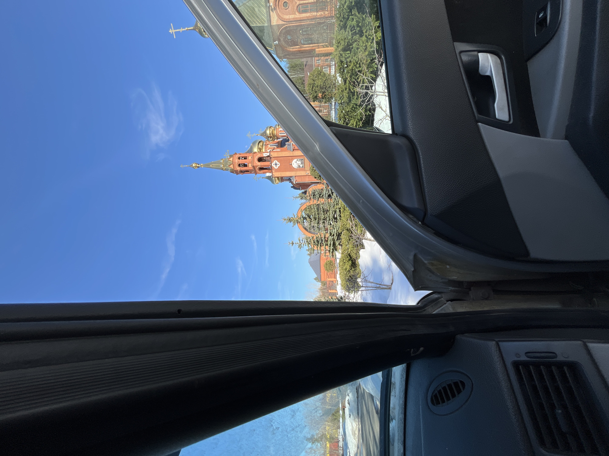 The image presents a view from inside a vehicle, looking outwards through an open driver's side door and a portion of the windshield. The interior of the vehicle shows sections of a dark grey dashboard with air vents, and the grey and black inner panel of the open door, featuring a silver door handle and window controls.
The external environment is centered on the Annunciation Cathedral (Благовещенский собор) in Chaikovsky, Perm Krai, Russia. This prominent structure is constructed from red brick, featuring multiple golden onion domes and a tall, multi-tiered bell tower crowned with a golden spire and a cross. A mosaic icon is visible on the cathedral's facade. The grounds surrounding the cathedral contain evergreen trees and patches of snow, indicating a winter or early spring season. The sky above is clear and blue with minimal cloud cover. No individuals are visible in the scene.