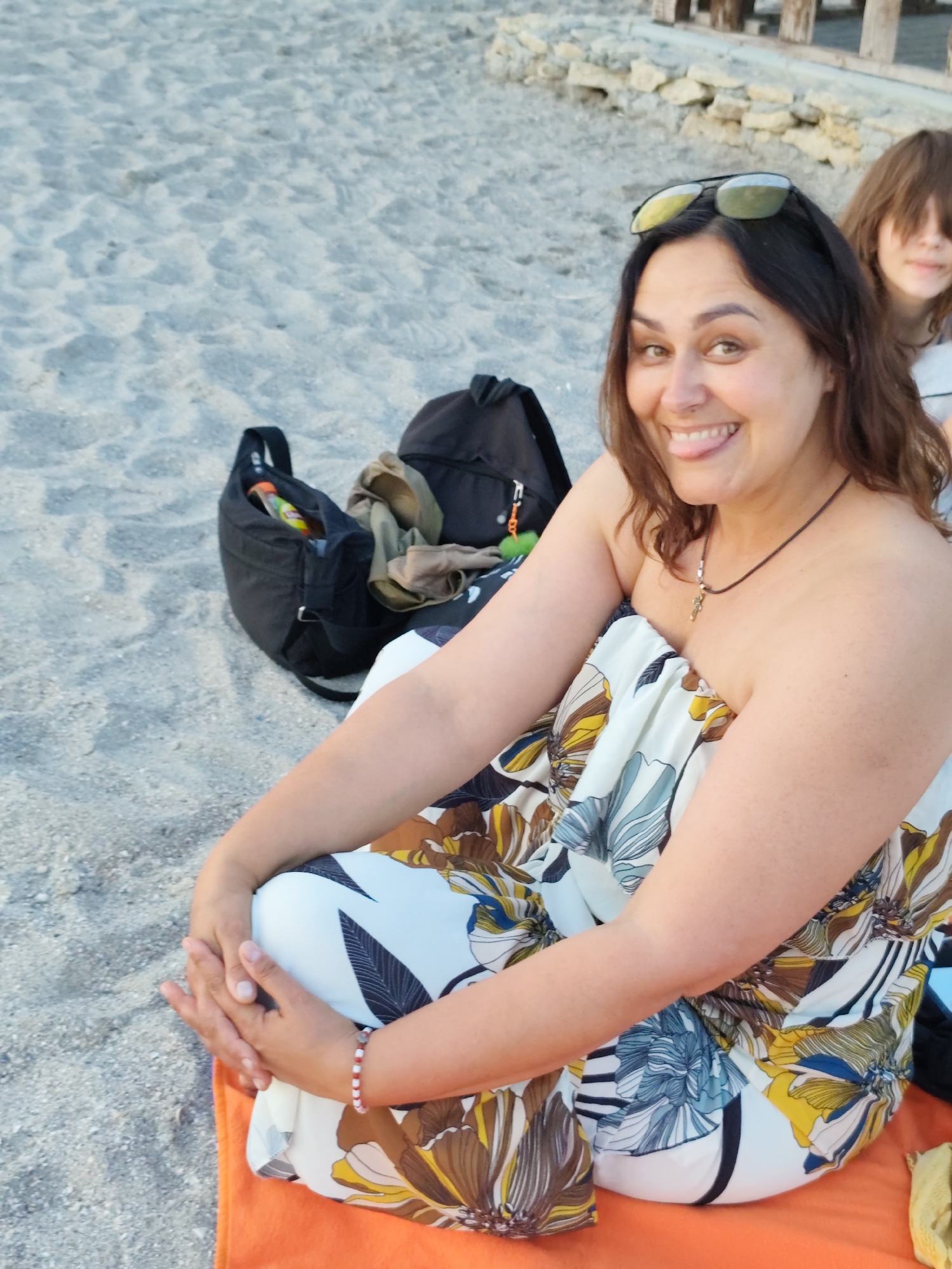 This media file captures a candid moment of two people on a sandy beach, likely during the late afternoon or early evening given the soft lighting. A woman in the foreground is the primary subject, sitting cross-legged on an orange towel, wearing a strapless dress with a floral pattern featuring brown, yellow, and blue motifs. She has dark hair, sunglasses perched on her head, and is looking at the camera with a playful expression, sticking out her tongue. A thin necklace with a cross pendant is visible around her neck, and she wears a beaded bracelet on her right wrist.
Behind her, a younger person with brown hair is partially visible, also appearing to be on the beach. Two backpacks are placed on the sand nearby, one black and the other a darker color, with a smaller bag containing an orange and yellow item peeking out from the black backpack. The background shows a sandy beach with scattered rocks and wooden structures, suggesting a casual, outdoor setting. The overall mood is relaxed and joyful, with the woman's expression conveying happiness. There is no visible text in the image.