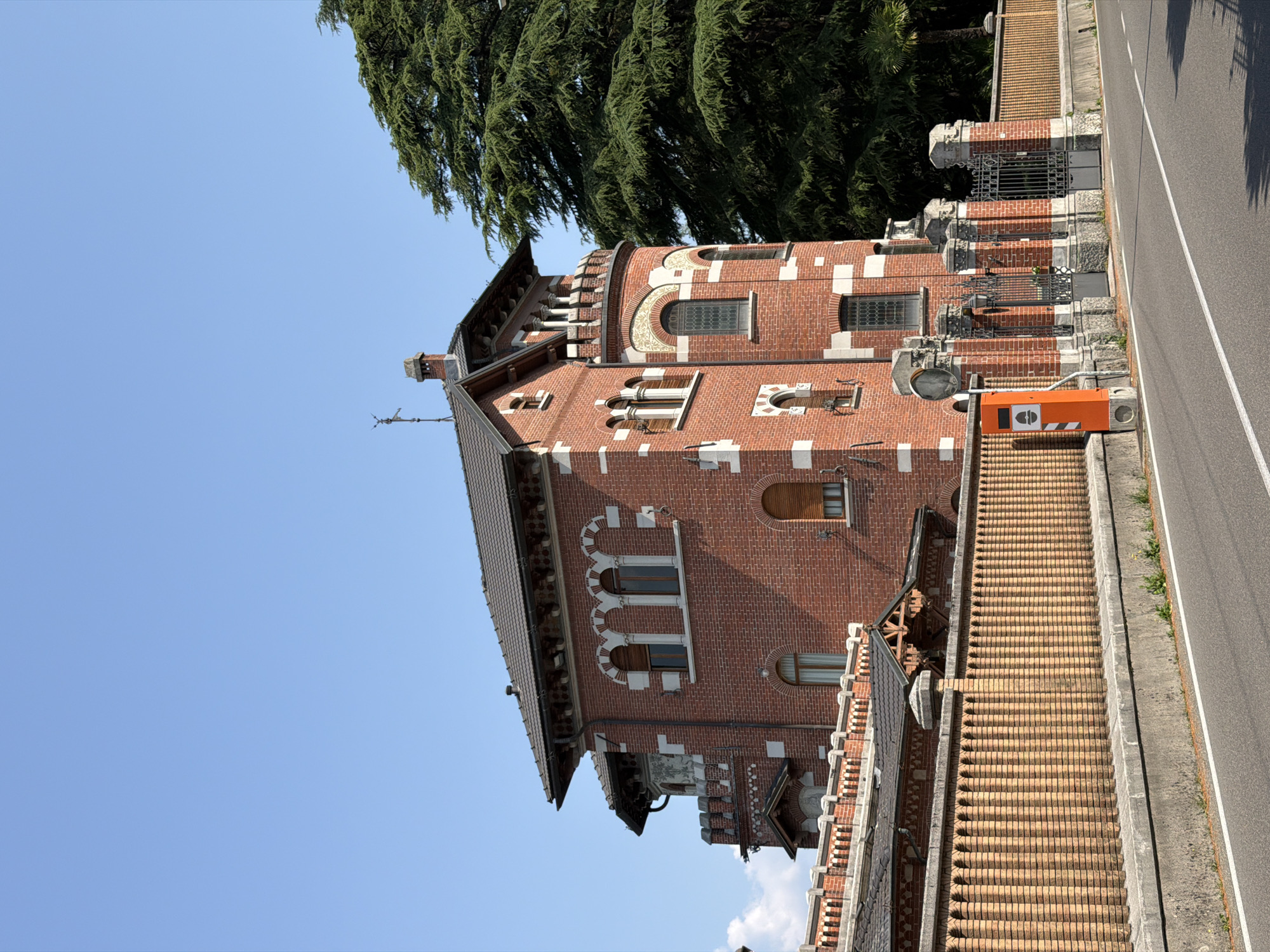 A bright, clear day with a cerulean sky over Lierna, Italy. The image showcases a distinctive red brick building with white accents, featuring multiple arched windows and decorative elements. A tall, ornate brick fence with a stone cap runs along the foreground, separating the property from the asphalt road. To the right of the property, a large, lush green tree provides a natural contrast to the architecture. An orange electronic device, possibly a parking meter or information kiosk, stands near the gate. The overall scene depicts a well-maintained and architecturally interesting property on a sunny day.