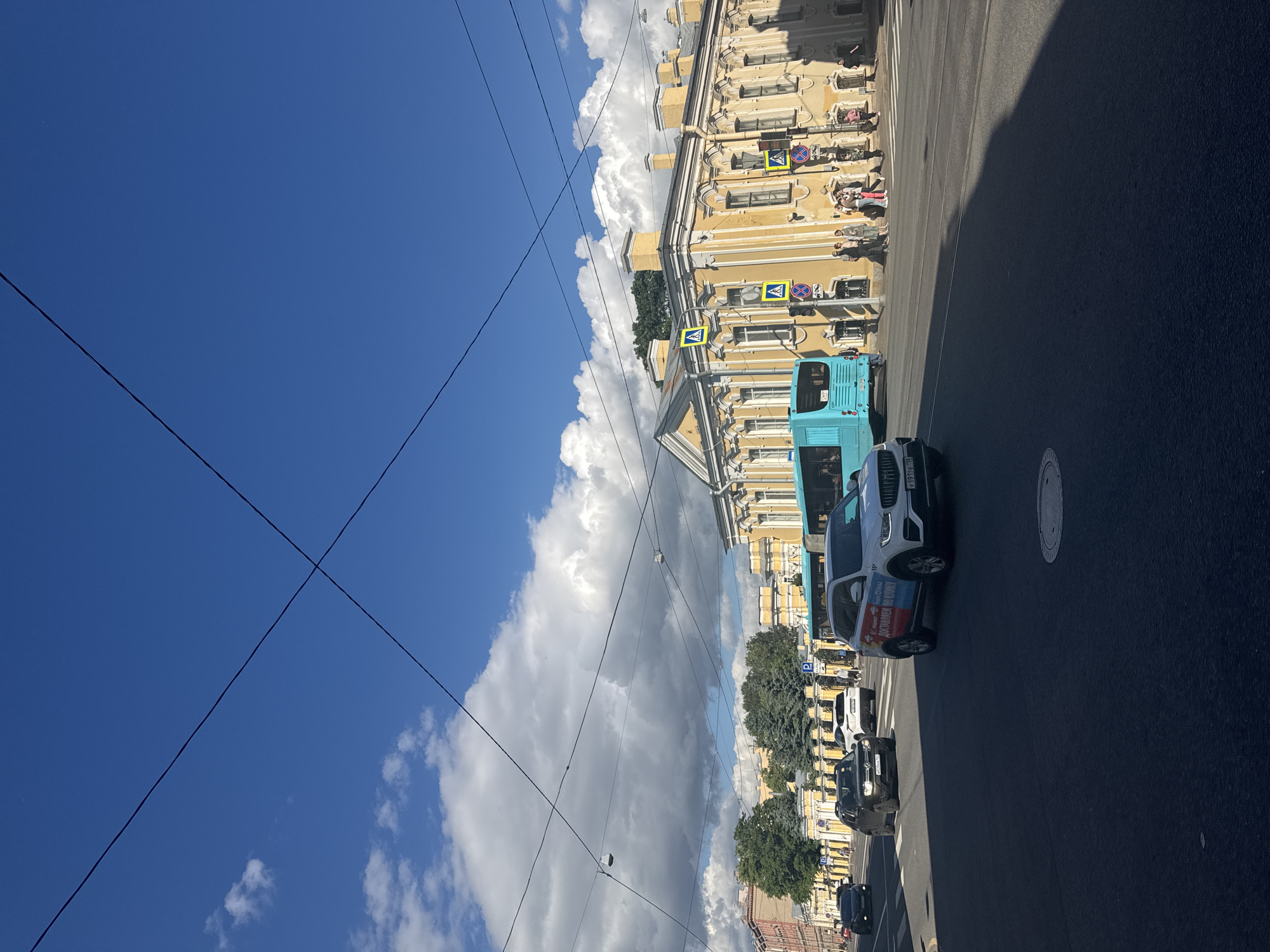 This image depicts a sunny day in a city, likely Saint Petersburg, Russia, with a bright blue sky and scattered white clouds. The scene is a street with buildings lining one side, characterized by their yellow facade and numerous windows. A teal-colored double-decker bus is parked on the street, alongside other vehicles, including a white SUV with red and blue lettering.
Several people are visible on the sidewalk in front of the buildings, appearing to be waiting or walking. There are traffic signs present, including pedestrian crossing signs. The time of day appears to be midday, given the strong shadows cast on the road and the brightness of the sky. Overhead power lines crisscross the frame, adding a layer of urban detail. The overall impression is of a typical urban street scene on a pleasant day, with hints of local life and transportation.